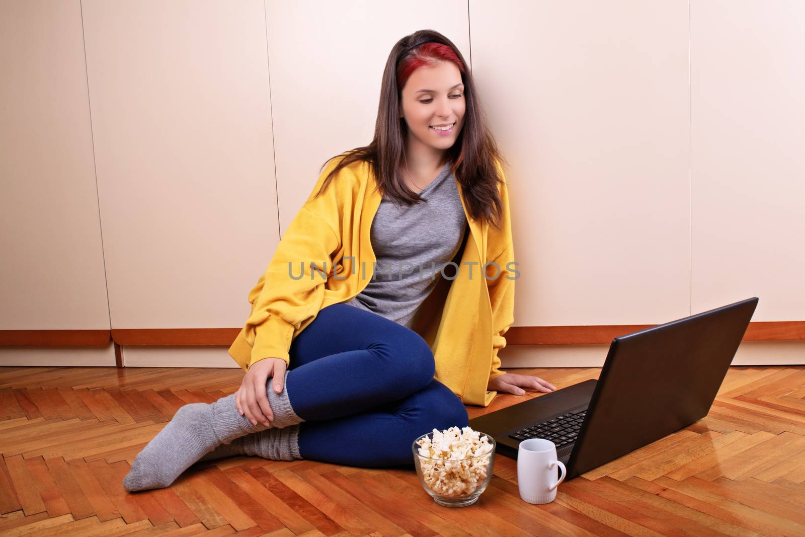 Beautiful smiling young girl in casual clothes sitting on the floor of her bedroom, ready to watch a movie on her laptop with a bowl of popcorn and a cup of coffee or tea. Leisure concept.