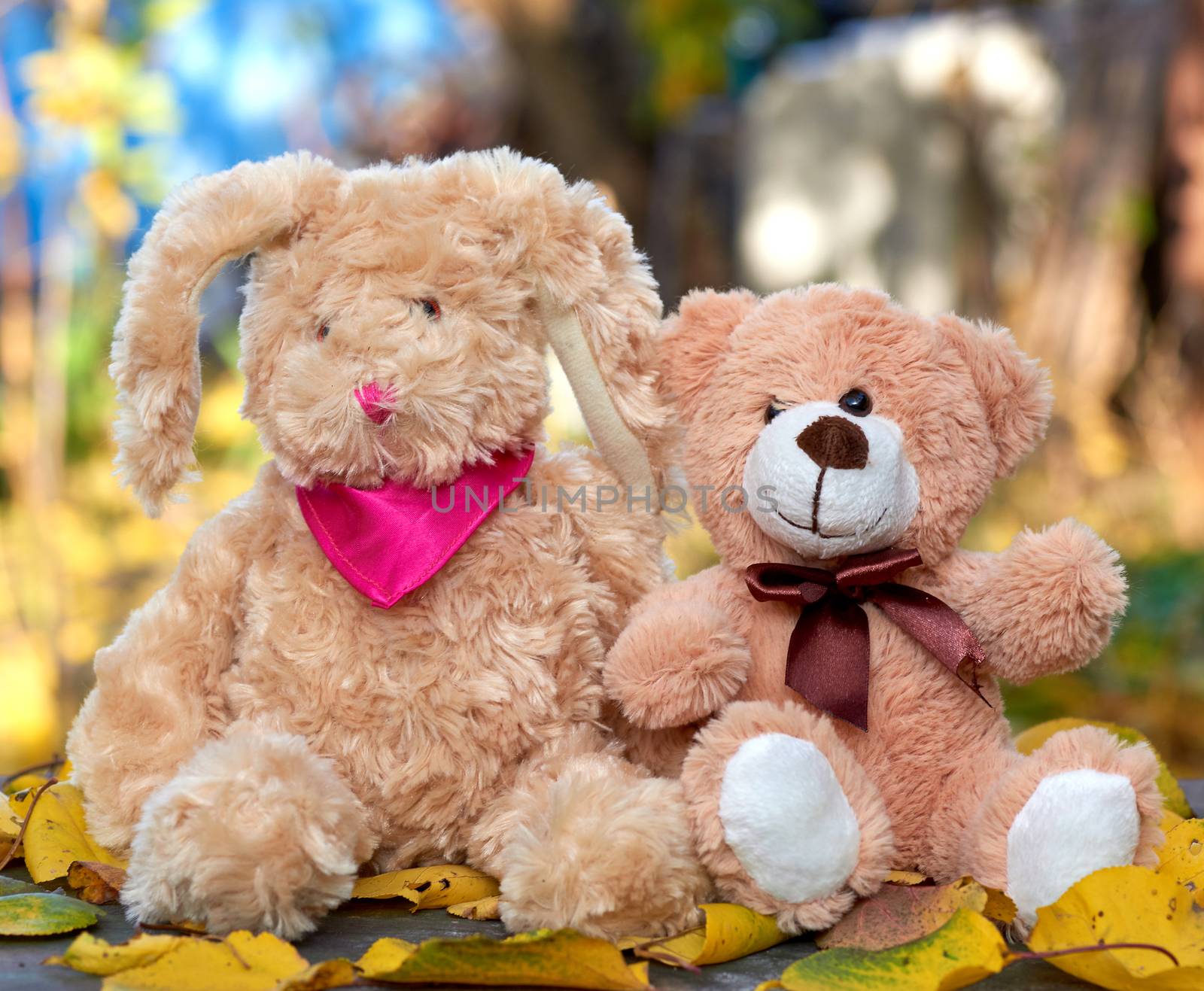little teddy bunny with long ears and a teddy bear are sitting in the middle of dry yellow leaves on an autumn day, blurred background behind, friendship concept
