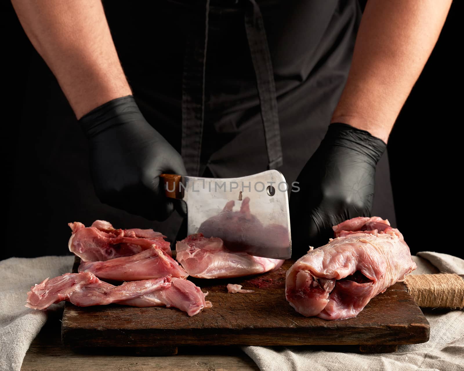 chef in black latex gloves holds a big knife and cuts into pieces raw rabbit meat on a brown wooden cutting board, cooking on a dark background