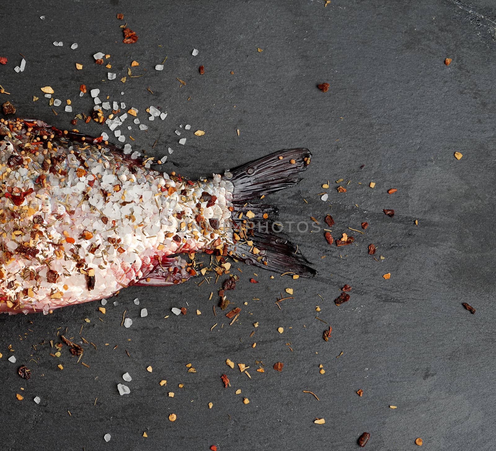 tail fresh crucian fish sprinkled with spices lies on a black slate board, top view