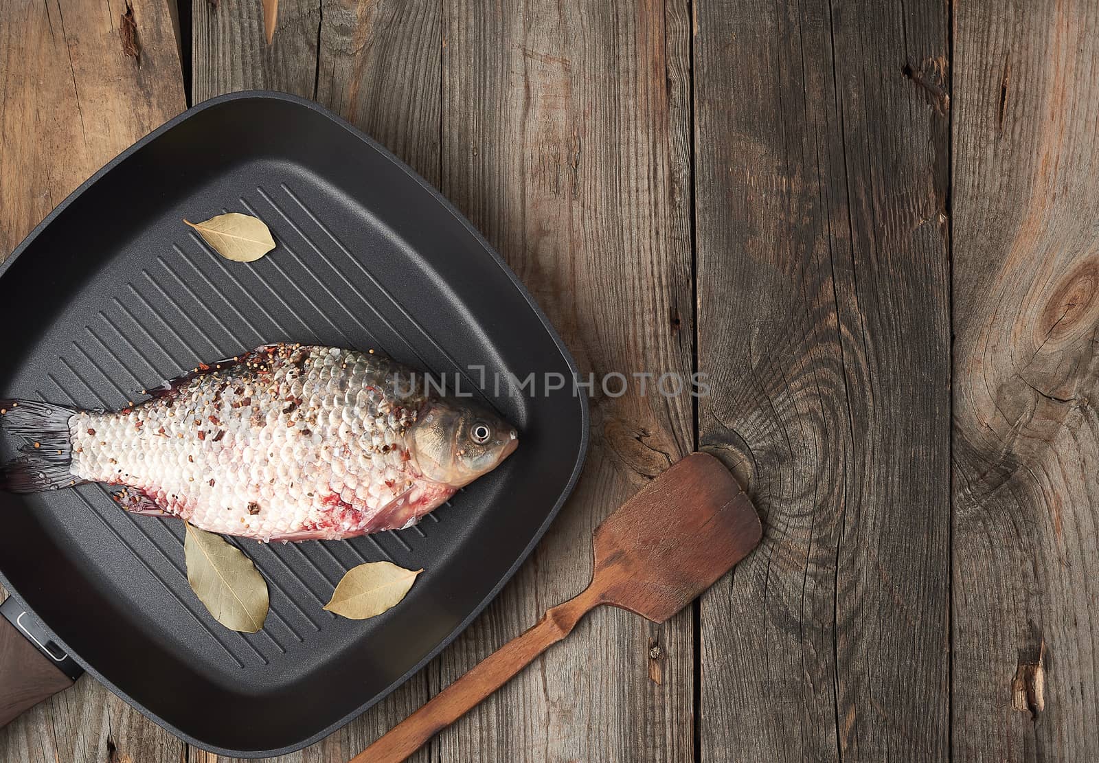 fresh crucian fish sprinkled with spices and lies in a black square pan, gray wooden table from boards, top view, copy space