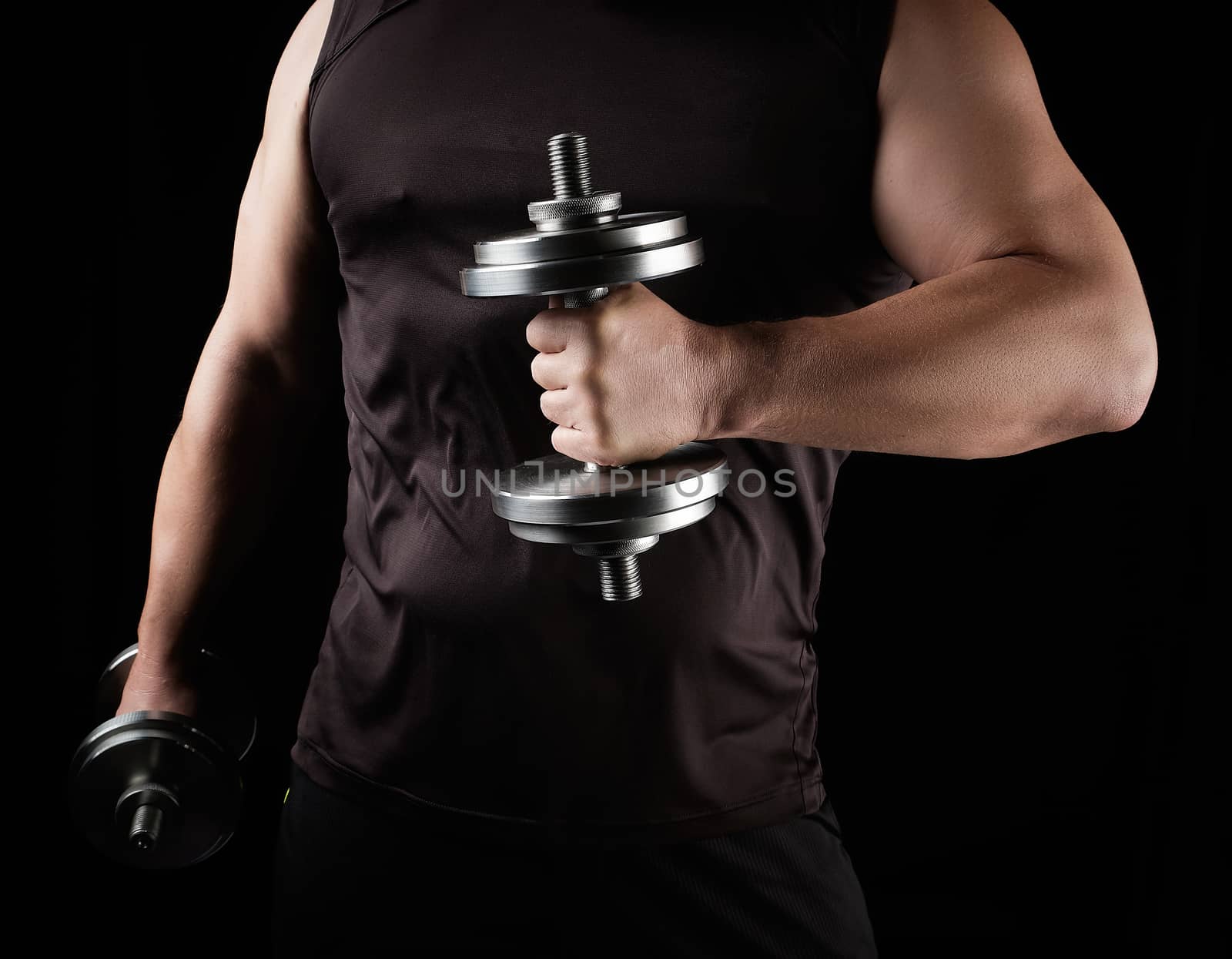 man in black clothes holds steel dumbbells in his hands, his muscles are tense, low key, sports background