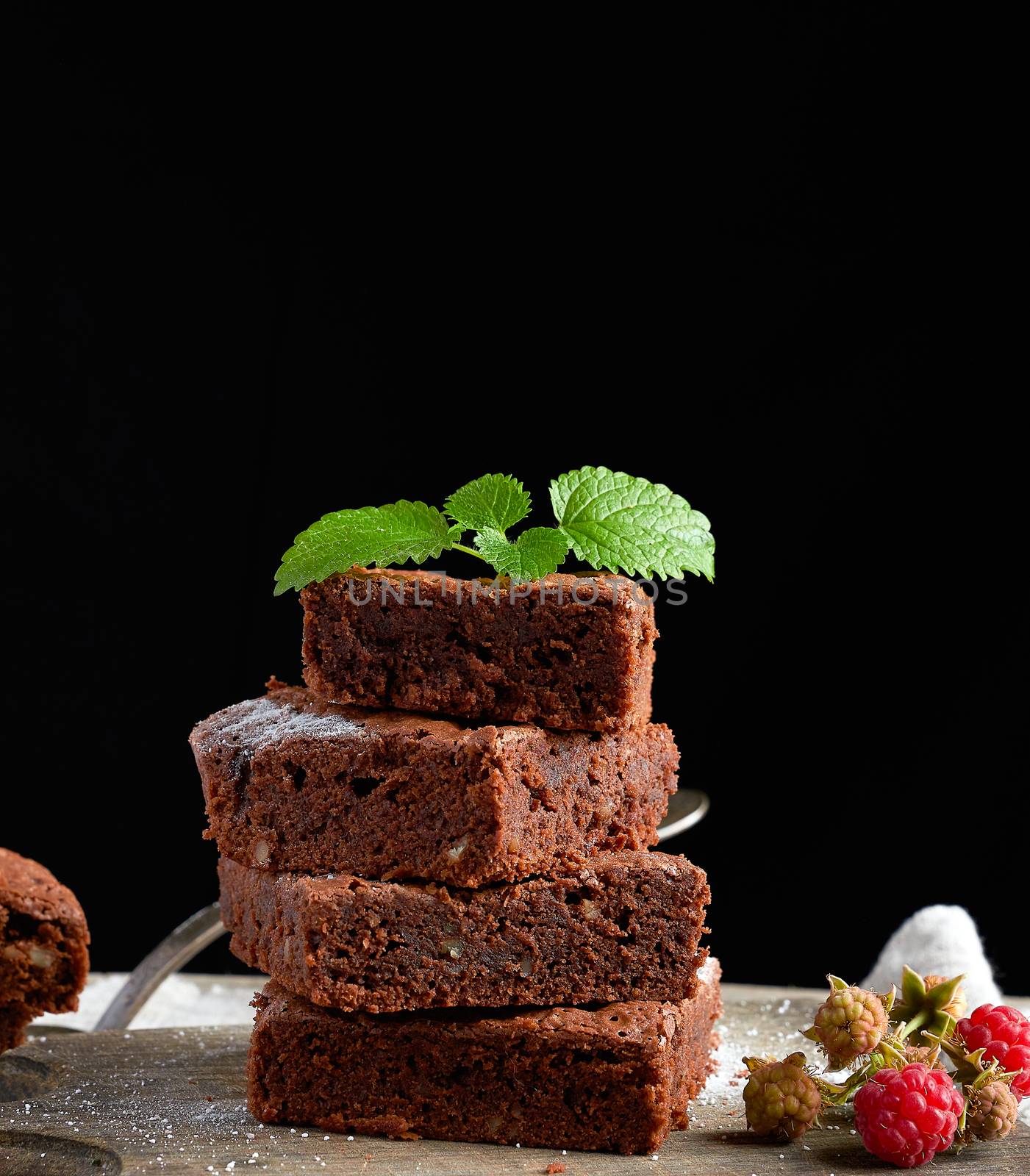 stack of baked square pieces of chocolate brownie cake on brown wooden cutting board, black background