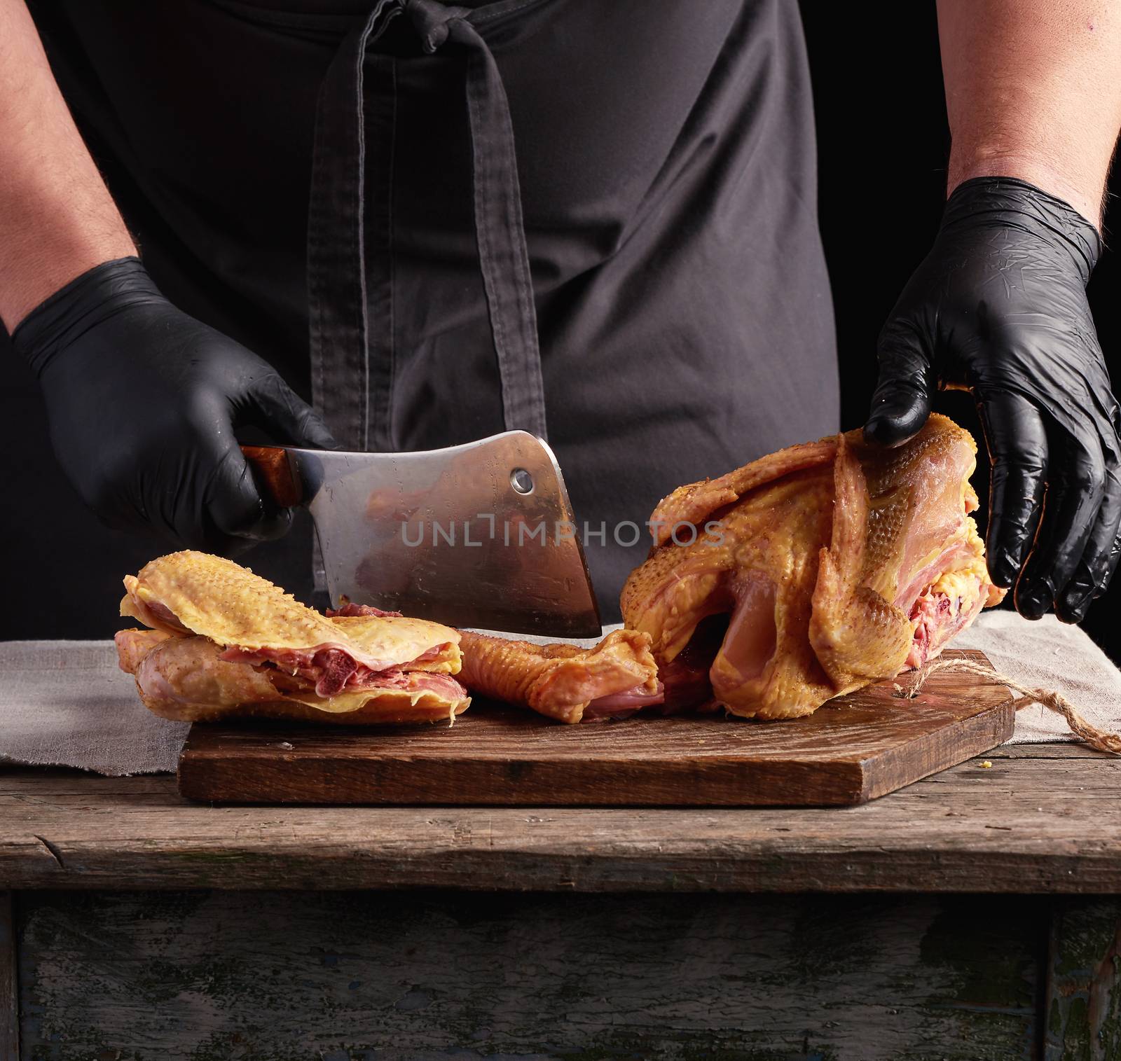 chef in black uniform and latex gloves chopping raw chicken into pieces on a brown wooden board, dark background, cooking process