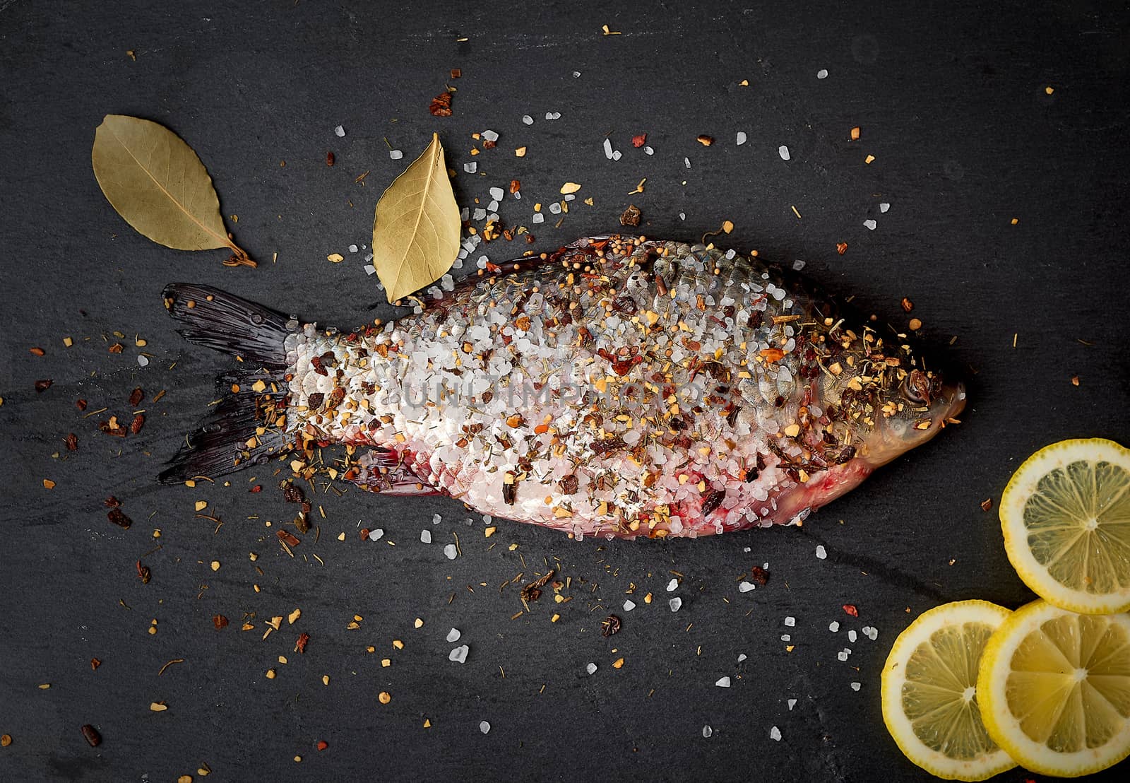 one fresh crucian fish sprinkled with spices lies on a black slate board, top view