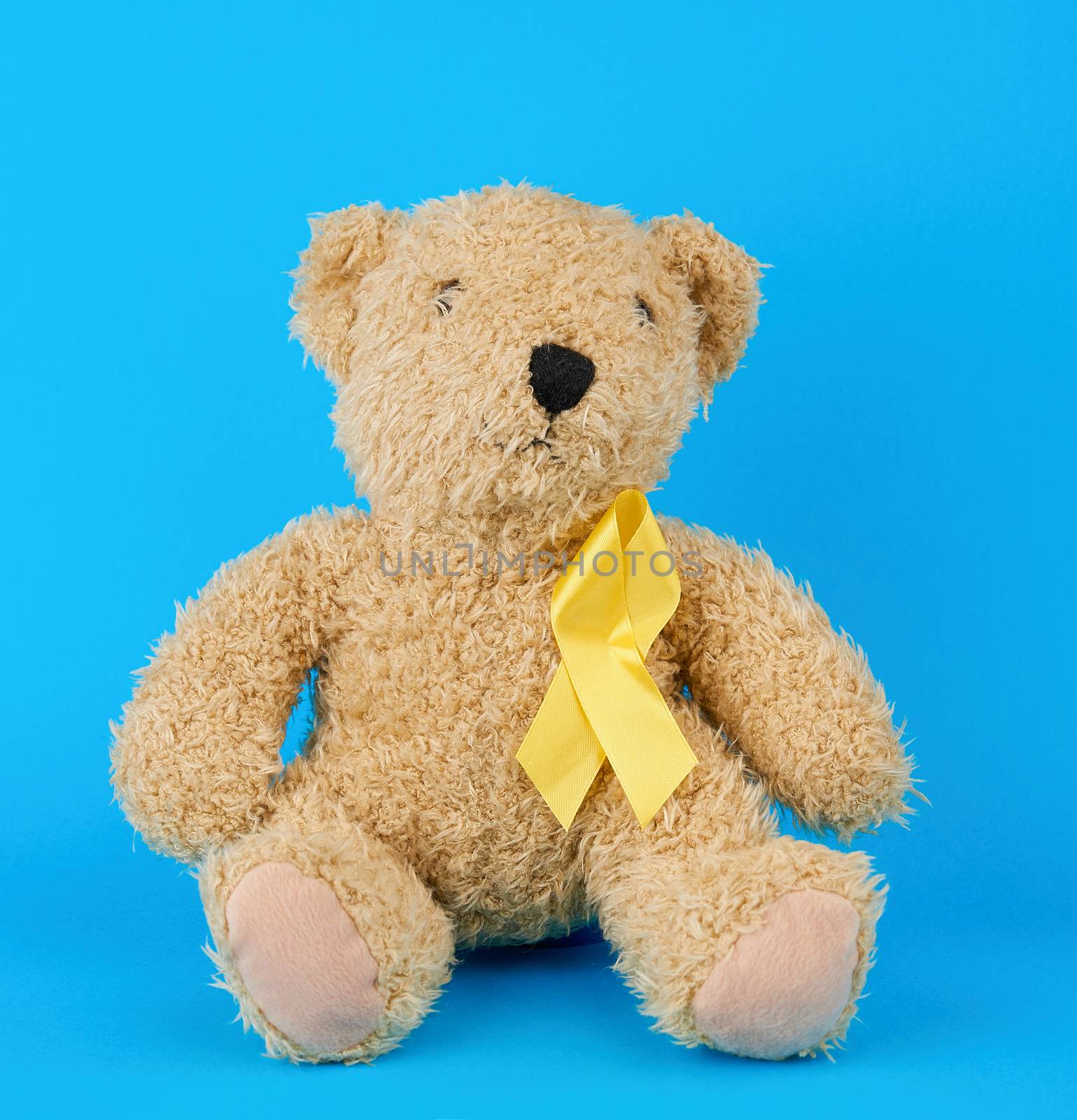 brown teddy bear sits and holds in his paw a yellow silk ribbon on a blue background, concept of the fight against childhood cancer. problem of suicides and their prevention