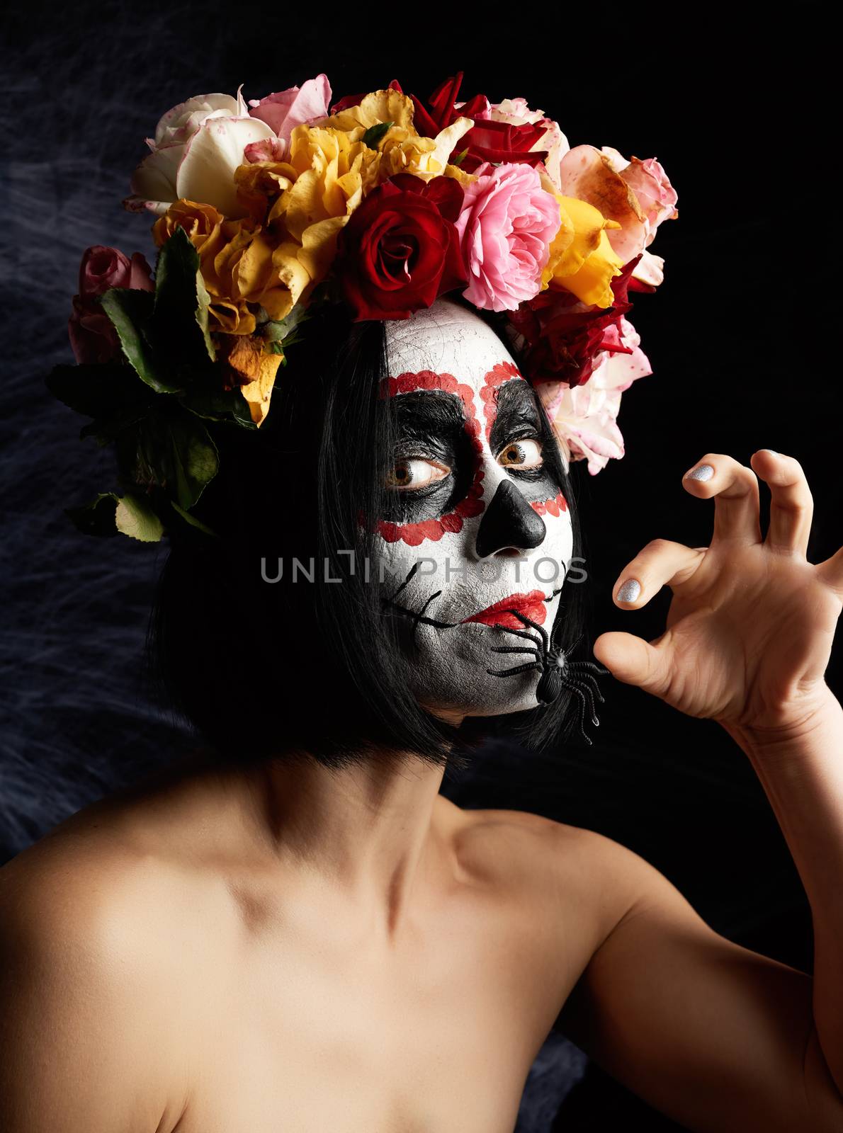 girl with black hair is dressed in a wreath of multi-colored roses and makeup is made on her face Sugar skull to the day of the Dead. Woman looking at the camera, black spider in mouth 