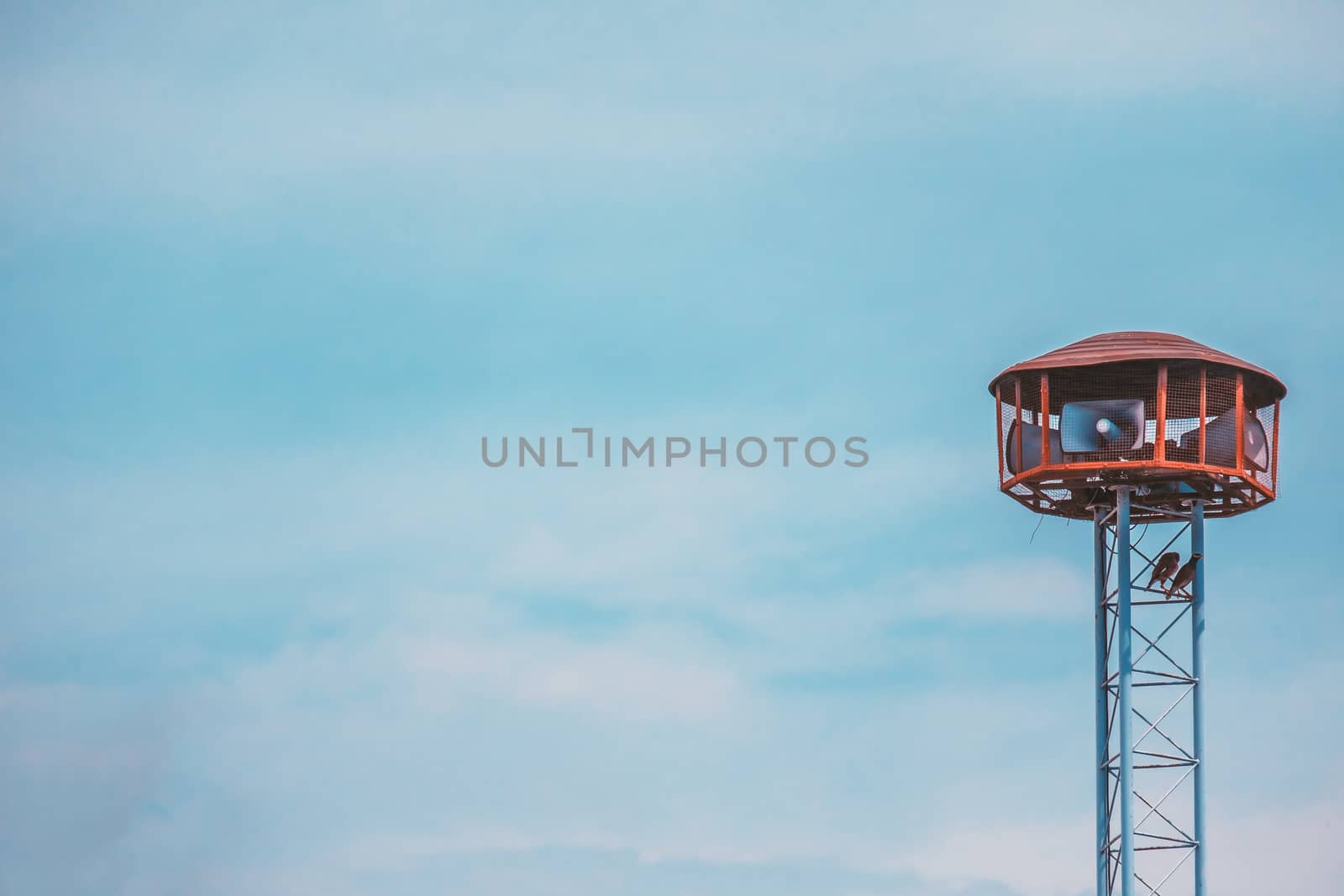 Public speaker tower with two birds perching  on against blue sky background and added retro color filter