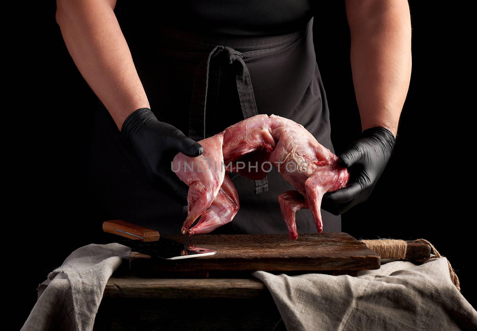 chef in black latex gloves holds a whole rabbit carcass over a brown cutting board, meat cooking process, dark background