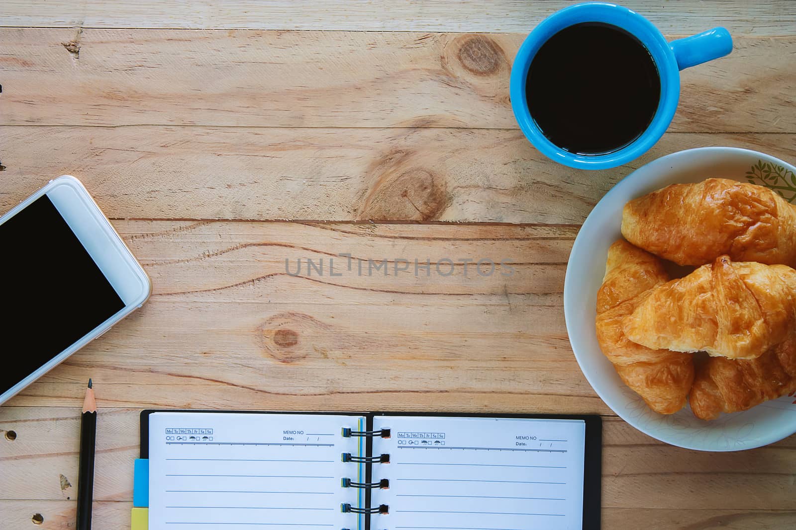 Top view of notebook, a cup of coffee, mini croissant bread, mobile smartphone,and pencil on the wooden table, space for thinking and writing with the snack time