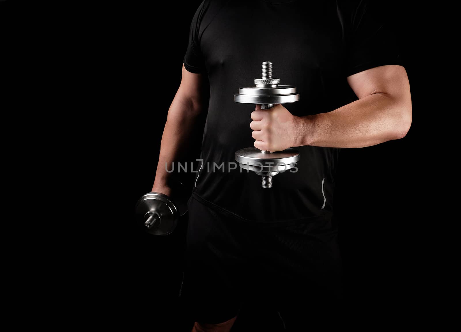 man in black clothes holds steel dumbbells in his hands, his muscles are tense, low key, sports background