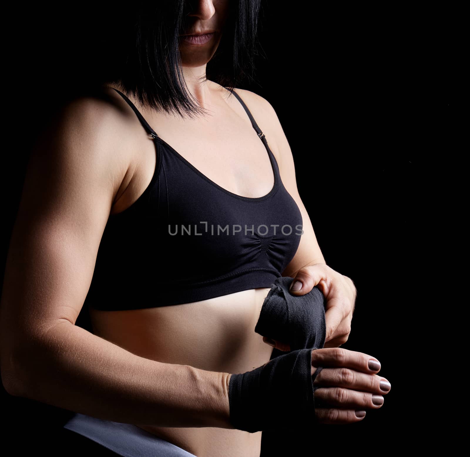 beautiful athletic girl with black hair rewinds her hand with a black elastic bandage before training, dark background