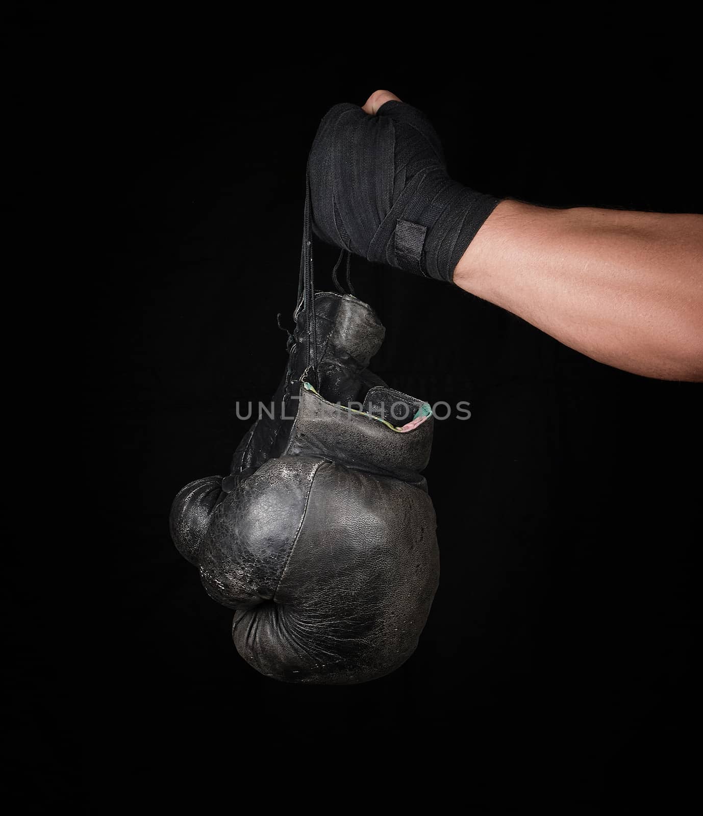 men's arm wrapped in a black elastic sports bandage holds pair old vintage leather boxing gloves, black background