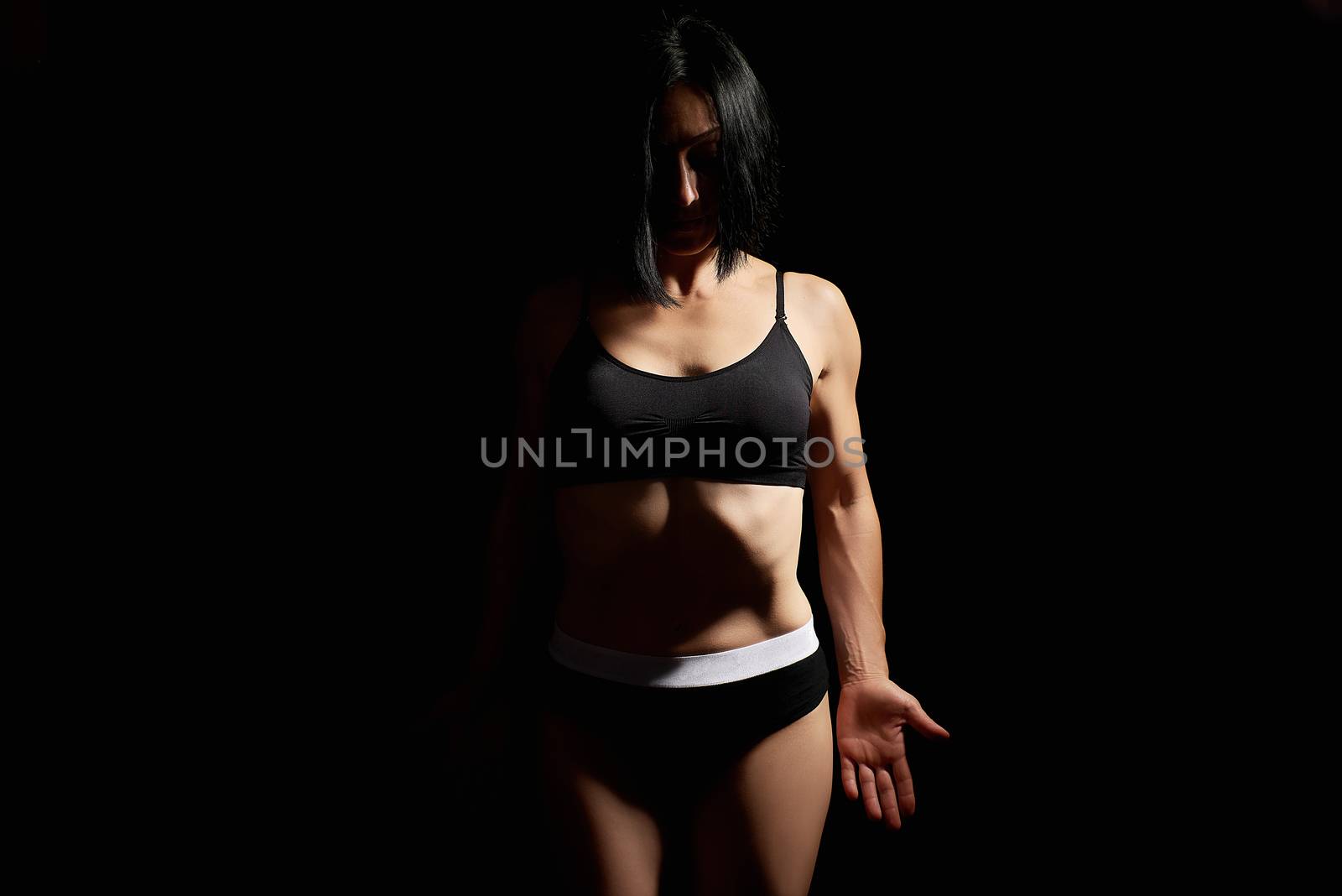 Adult girl with a sports figure in black bra and black shorts standing on a dark background, muscular body, black hair