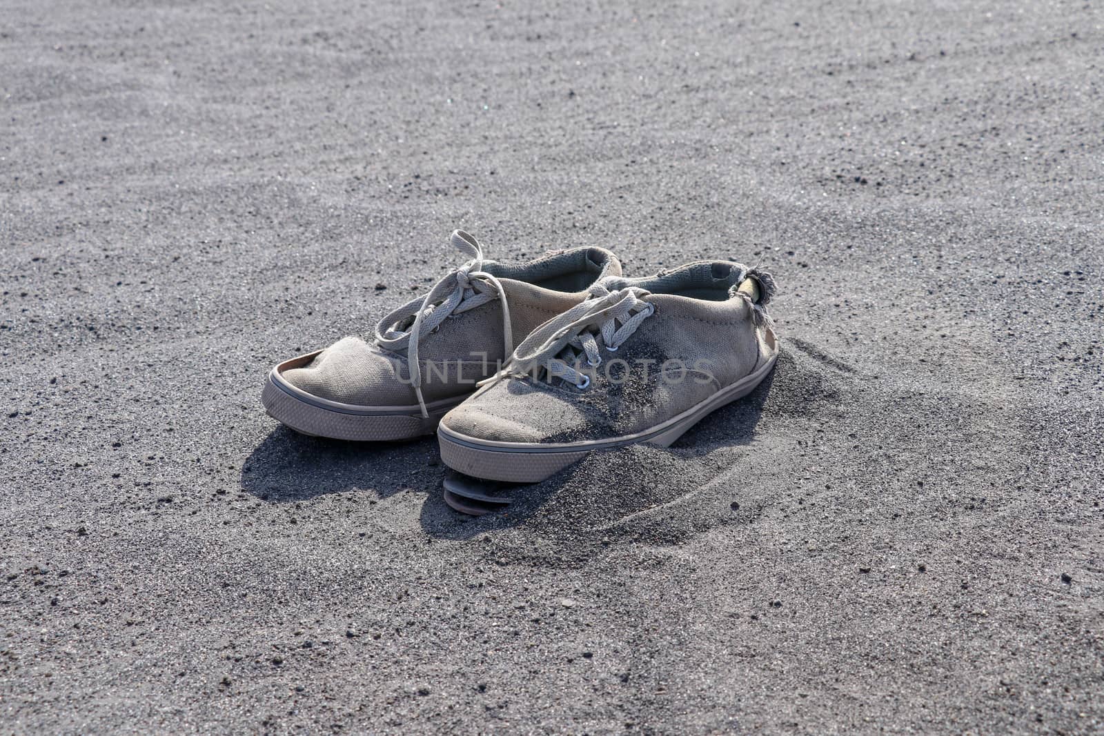 Old torn white sneakers on black volcanic sand. A pair of tie shoes with laces in volcanic ash. Discarded destroyed shoes.