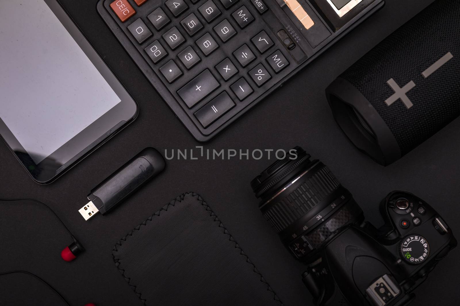 Top view of pitch black office desk with calculator, usb drive, earphones, dslr camera and wireless speaker. Minimal black design concept.