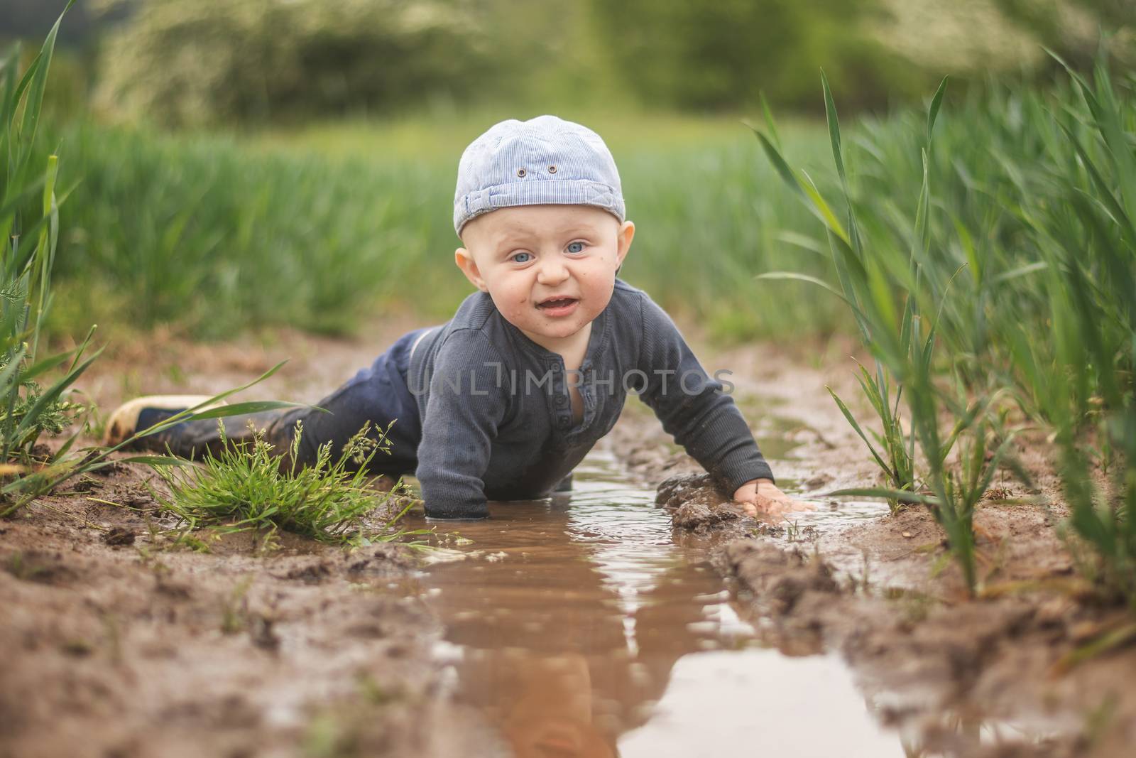 A white infant in a cap plays in a muddy pool. Children's discovery of the world. Looking into the camera.