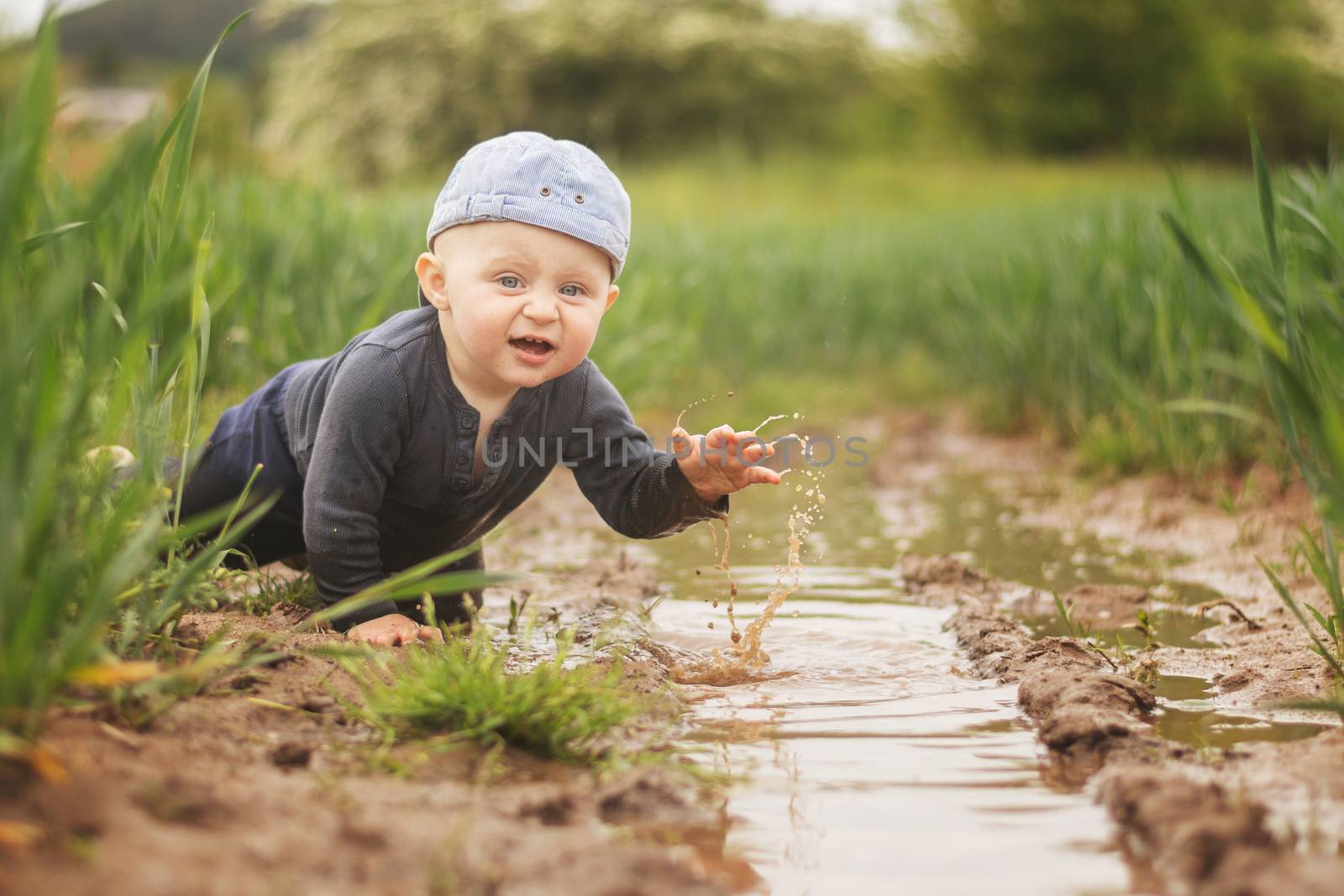 A Caucasian infant in a cap is playing in a muddy pool. Children's joy of freedom. Looking into the camera.