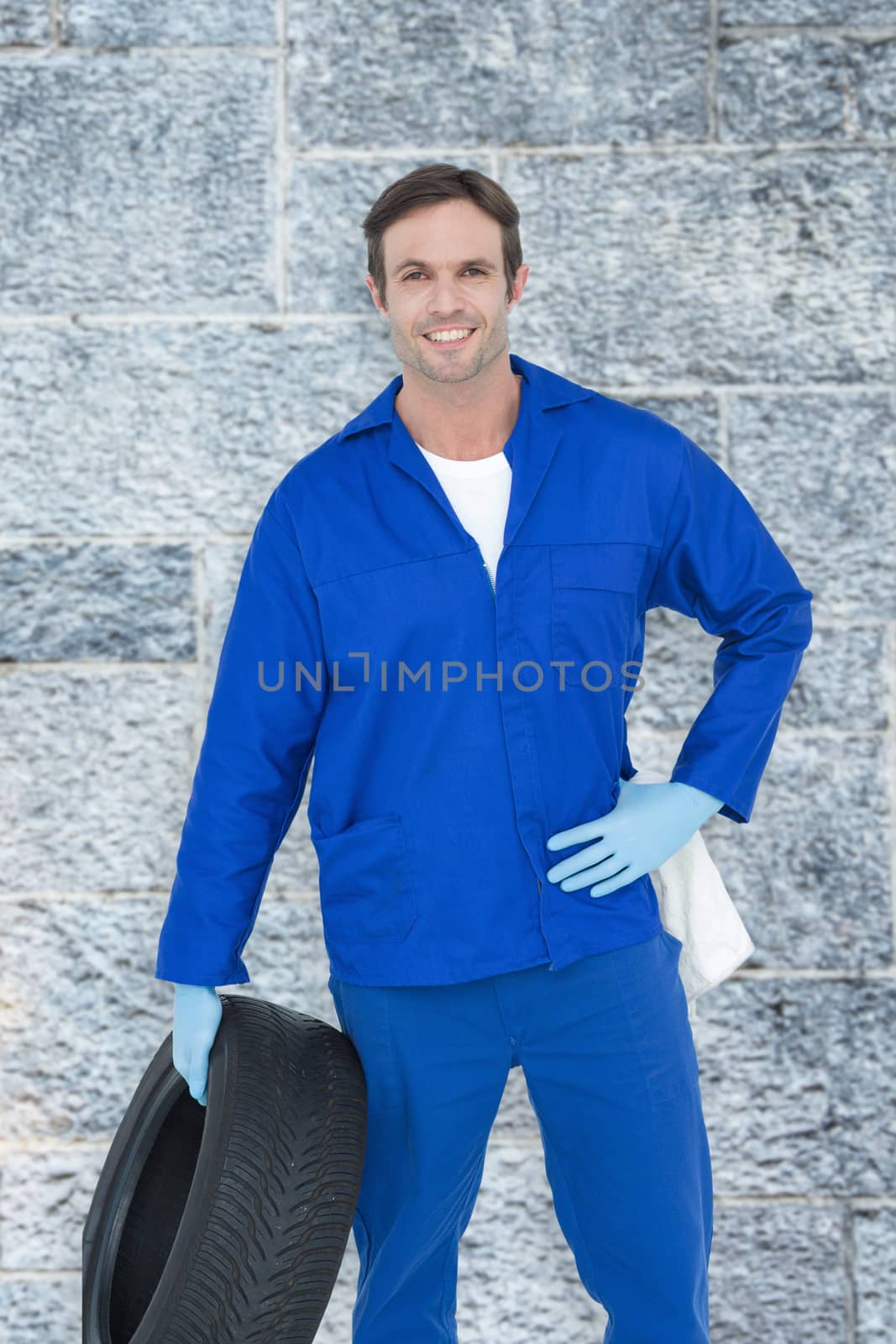 Mechanic holding tire while standing with hand on hip against grey