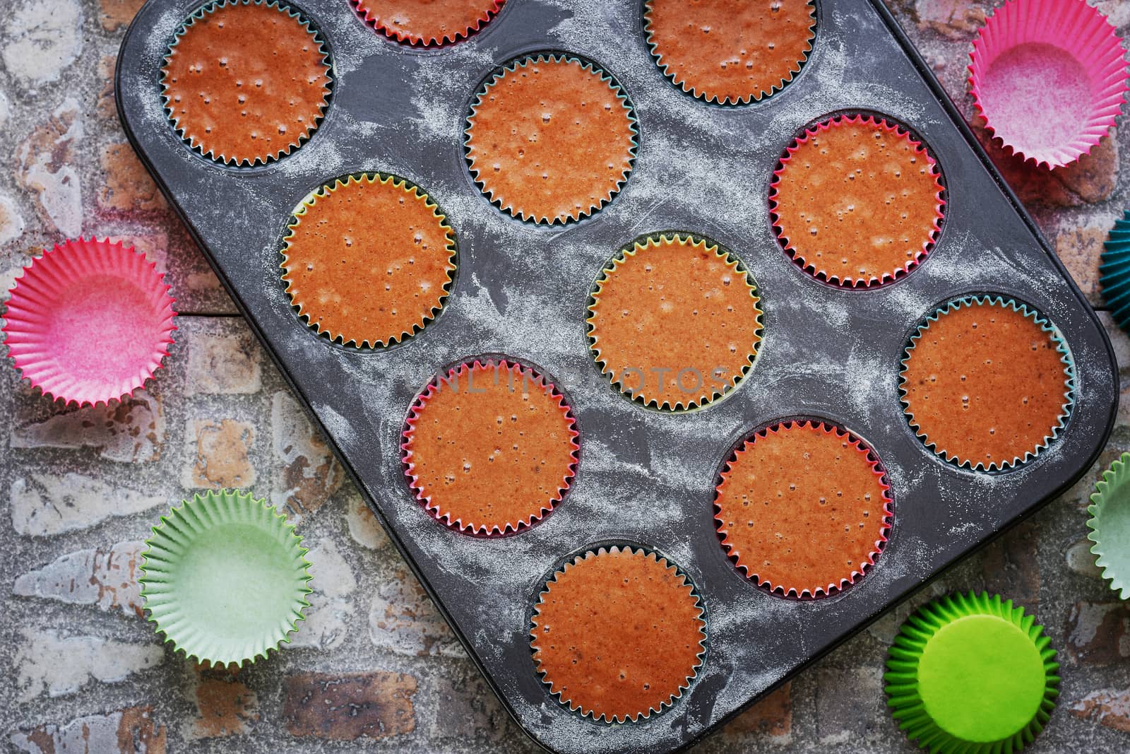 Baking tray for muffins with raw dough on earthenware background surrounded by paper colored baskets. View from above.