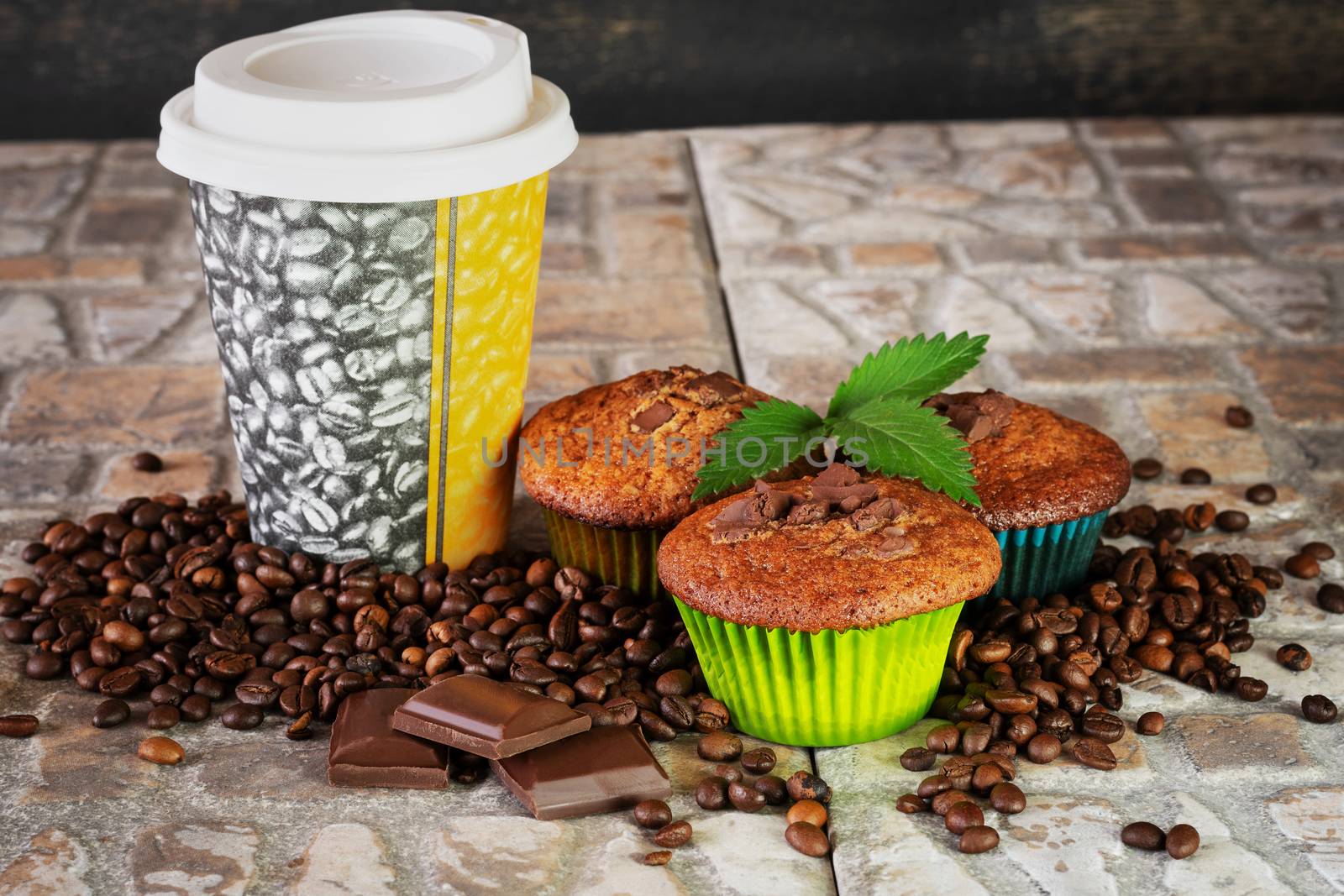 Muffins and coffee to go with green sprig, pieces of chocolate and coffee beans on stoneware background.