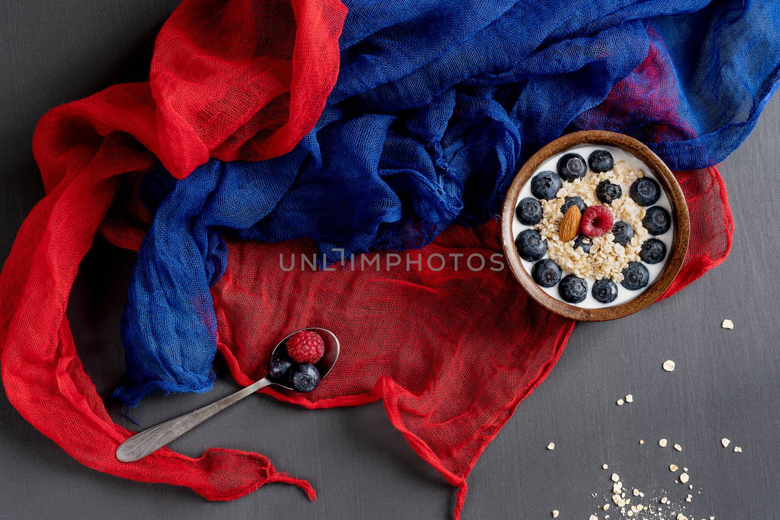 Healthy food. Bowl of oatmeal with blueberries, spoon, blue and red scarf on a gray table.