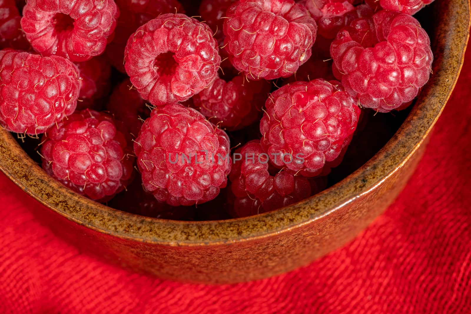 A clay bowl filled with red raspberries on a red scarf. Close up.