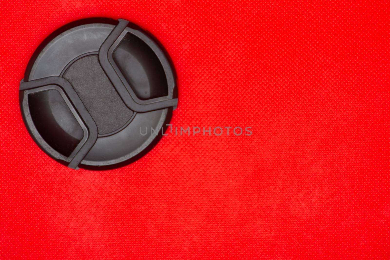 Close up of a black circle lens cap top left corner for DSLR camera lens on a rich red background.