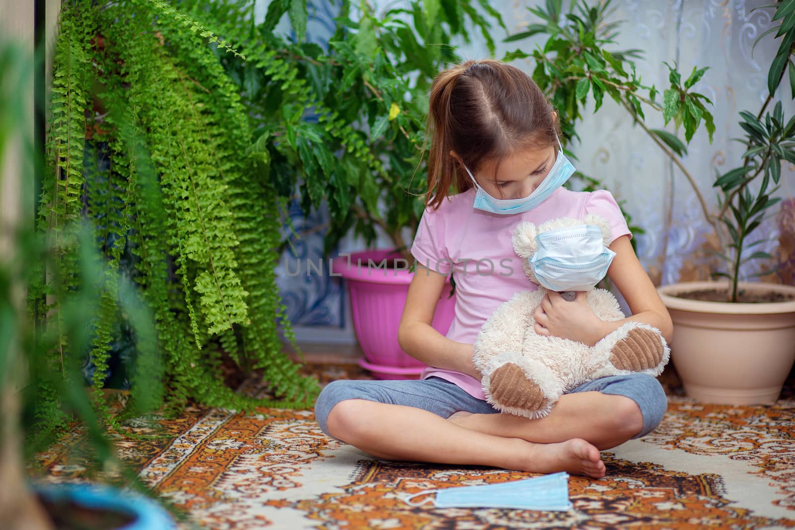 A girl in a medical mask sits at home during a Vieste epidemic with her teddy bear also in a mask