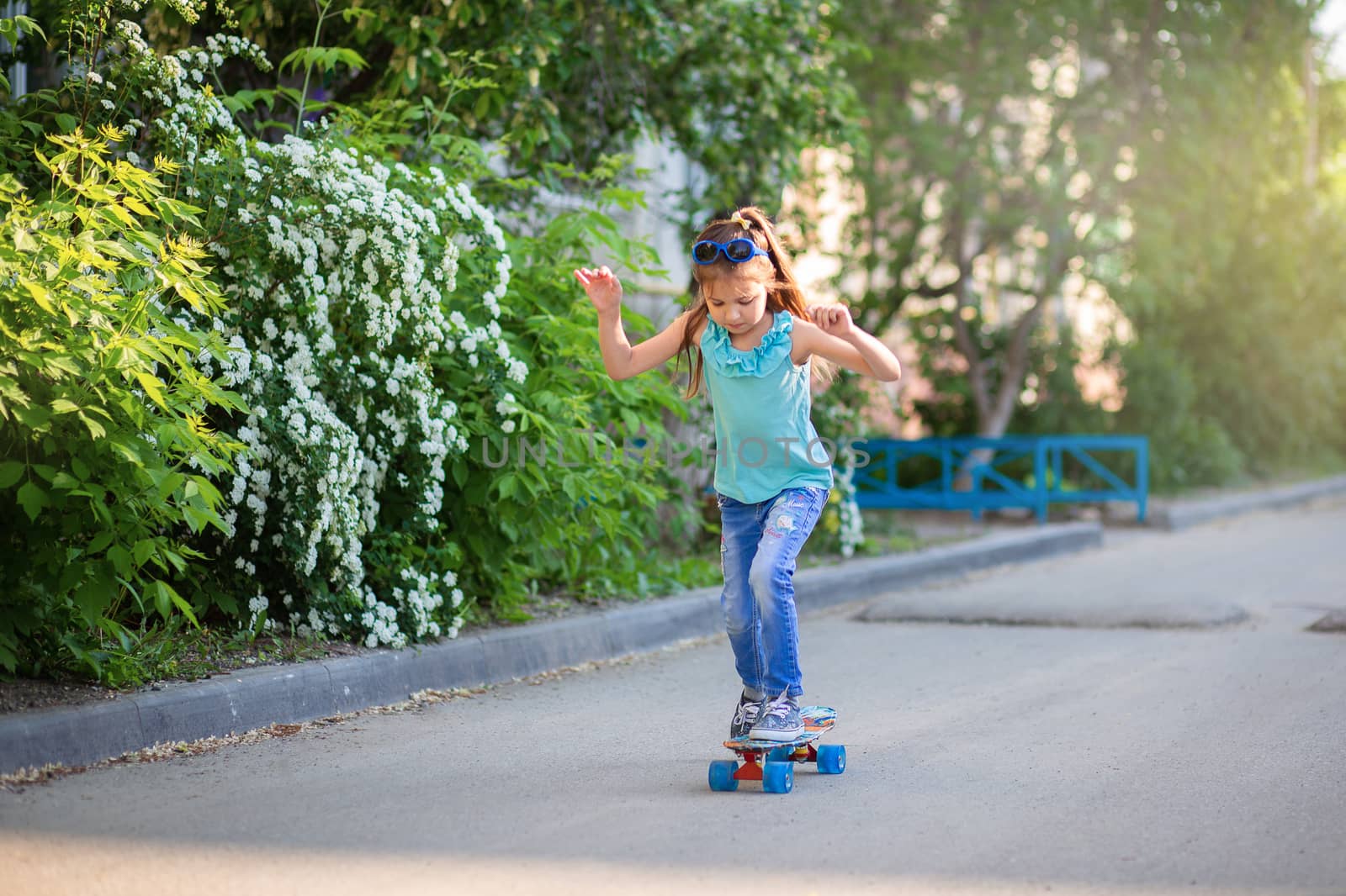 A little girl in jeans and sunglasses rides a skateboard around the yard in spring or summer. Children's sports at home