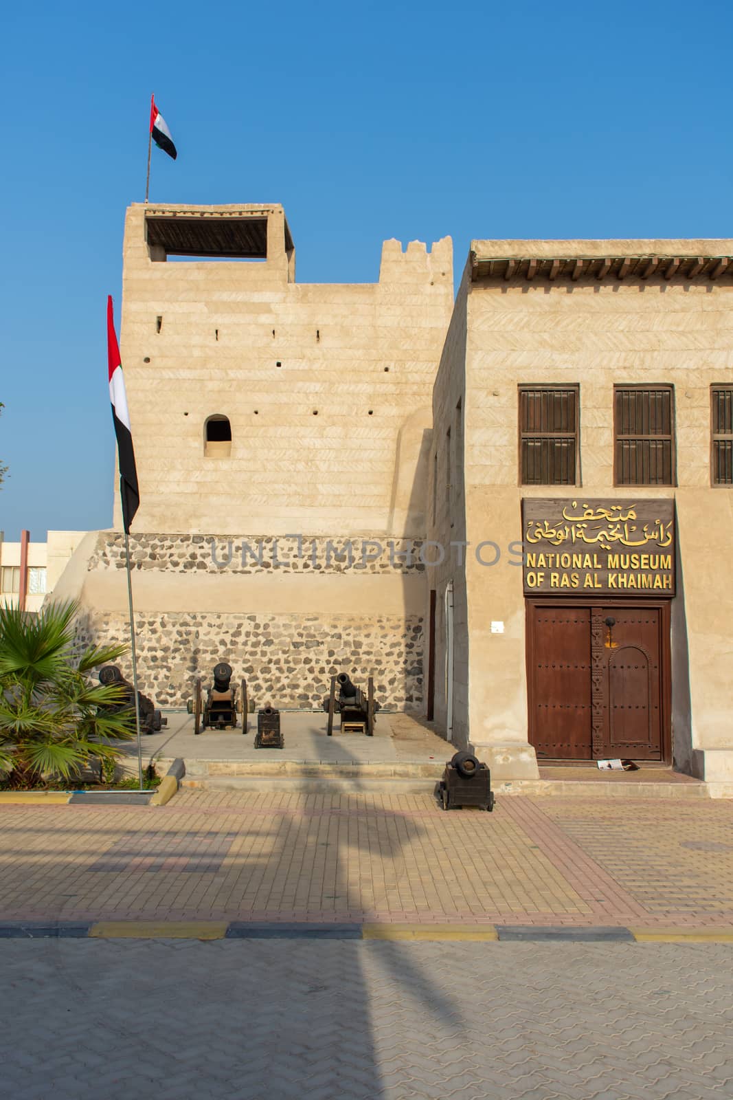 "Ras al Khaimah, Ras al Khaimah/United Arab Emirates - 9/28/2018: Portrait view of entrance of the Ras al Khaimah Museum in the morning sun with flags blowing."