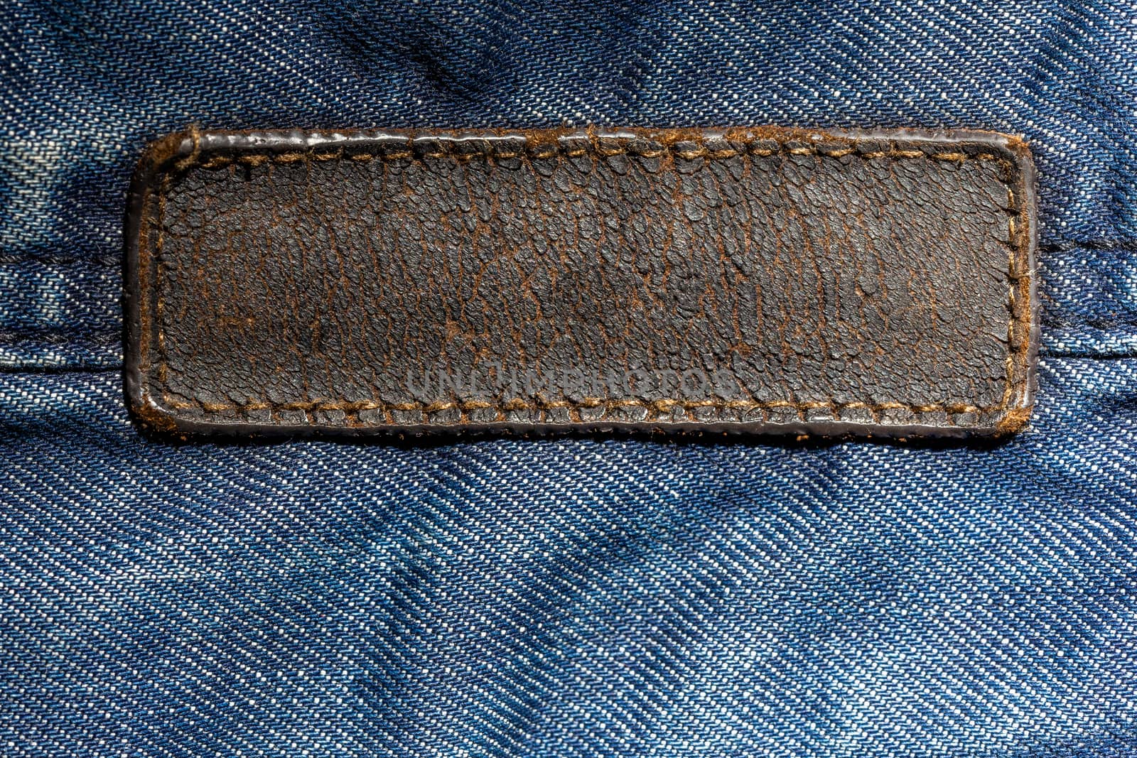 old weathered and cracked blank leather label on blue denim cloth - close-up with selective focus and partial blur. long tag