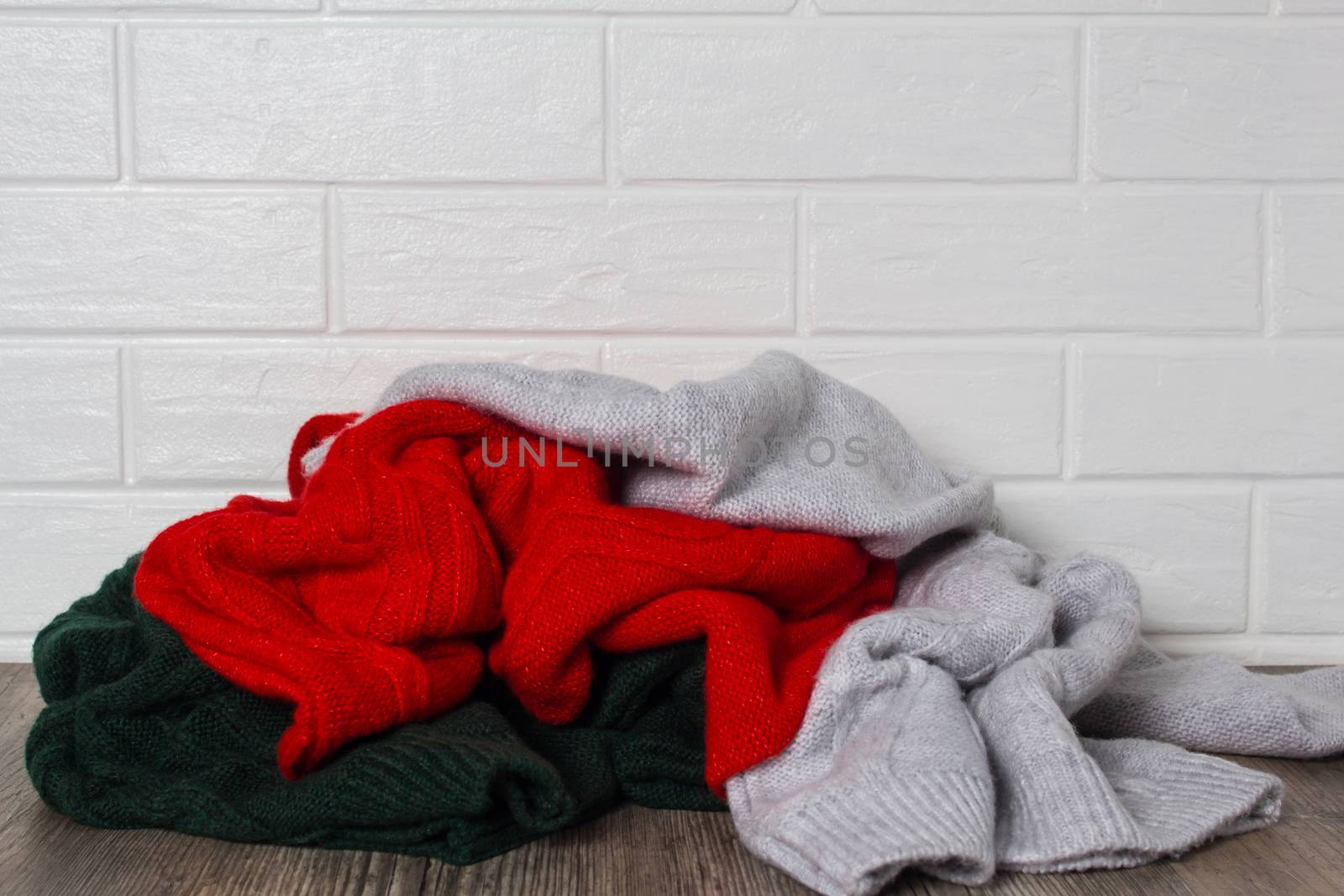 A pile of clothes against a white brick wall. Fashion, style. Sweaters in red, green and melange colors. Mess, home routine, storage. Wardrobe, hygge and knitwear.