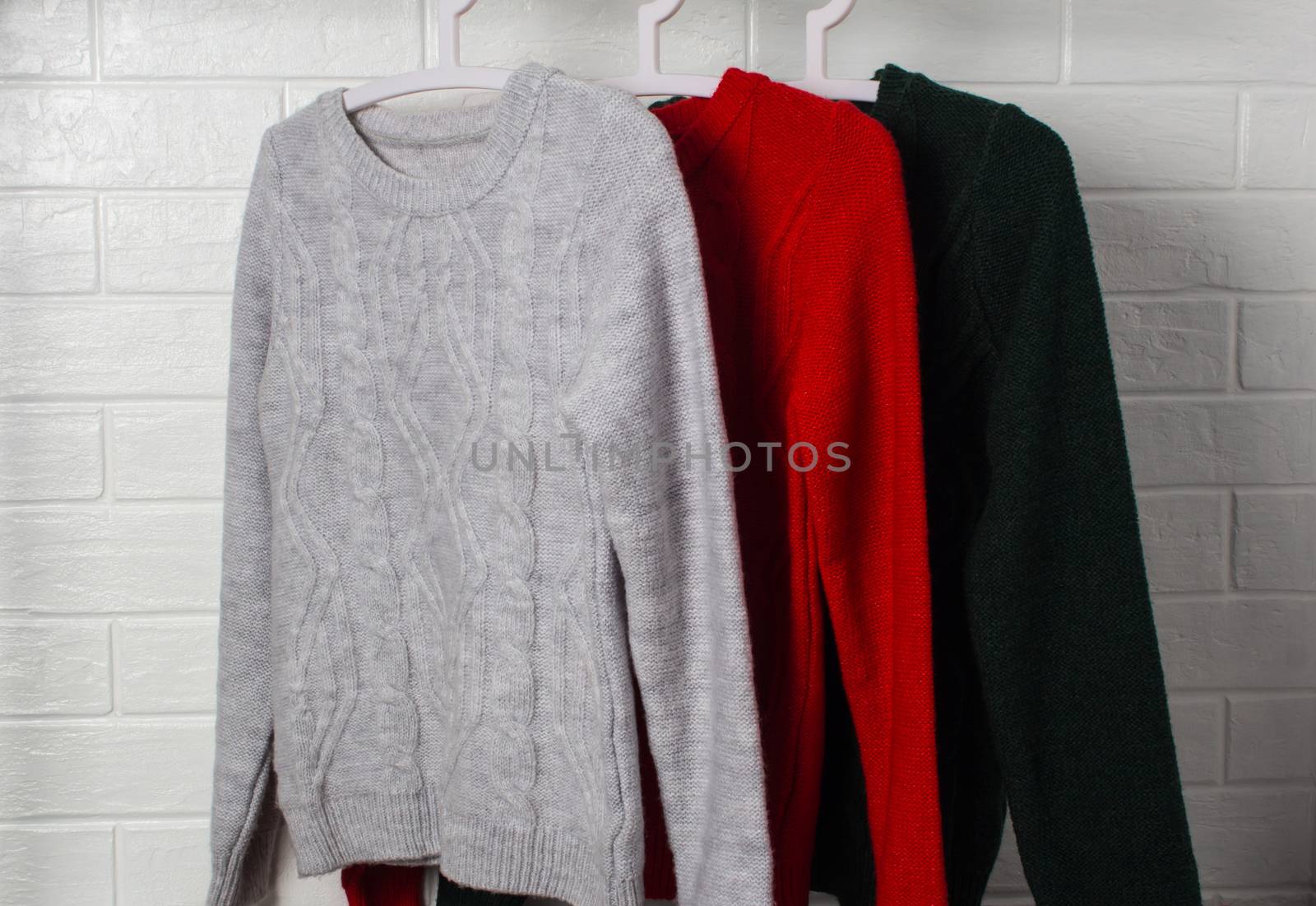 Clothes hang on hangers. Sweaters in red, green and melange colors. Fashion and style, showcase, store, concept. White brick wall, comfort and cleaning, design, hygge, soft and knitwear. Wardrobe.