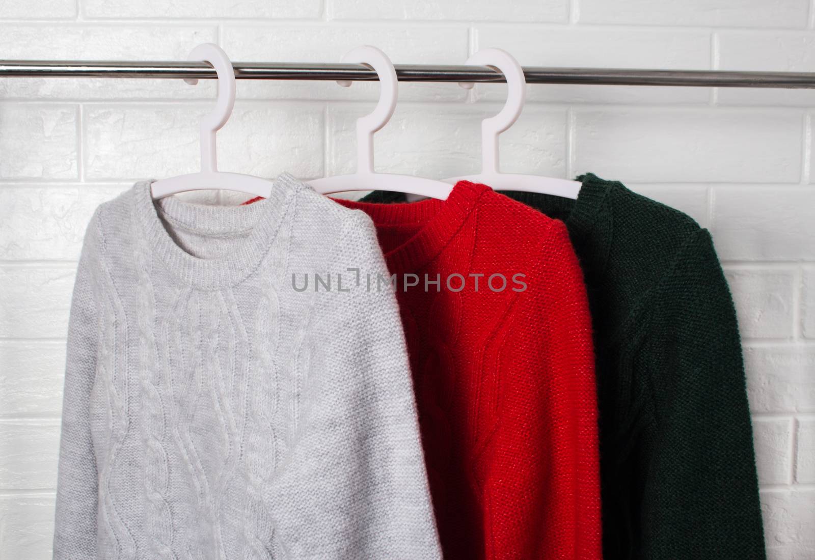 Clothes hang on hangers. Sweaters in red, green and melange colors. Fashion and style, showcase, store, concept. White brick wall, comfort and cleaning, design, hygge, soft and knitwear. Wardrobe.
