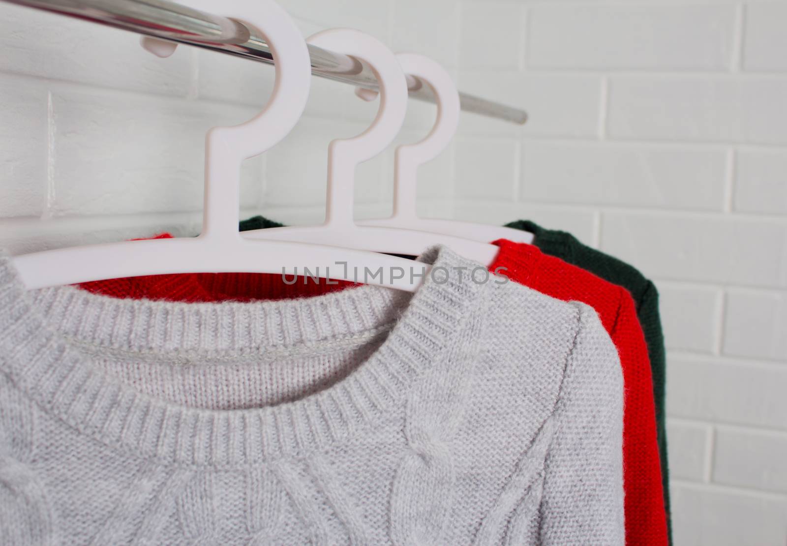 Clothes hang on hangers. Sweaters in red, green and melange colors. Fashion and style, showcase, store, concept. White brick wall, comfort and cleaning, design, hygge, wardrobe. and knitwear. Close up