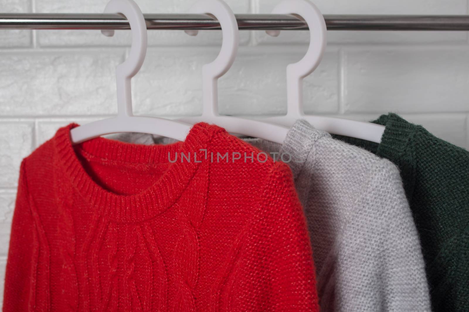 Clothes hang on hangers. Sweaters in red, green and melange colors. Fashion and style, showcase, store, concept. White brick wall, comfort and cleaning, design, hygge, soft and knitwear. Wardrobe.