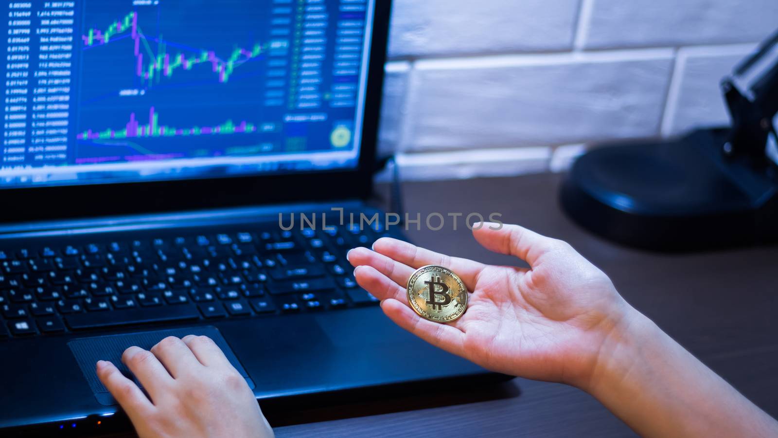 Bitcoin in hand against the background of trading charts. Cryptocurrency. A girl holds a Bitcoin coin sitting in front of a laptop analyzing a trader's price chart on the exchange or Forex.