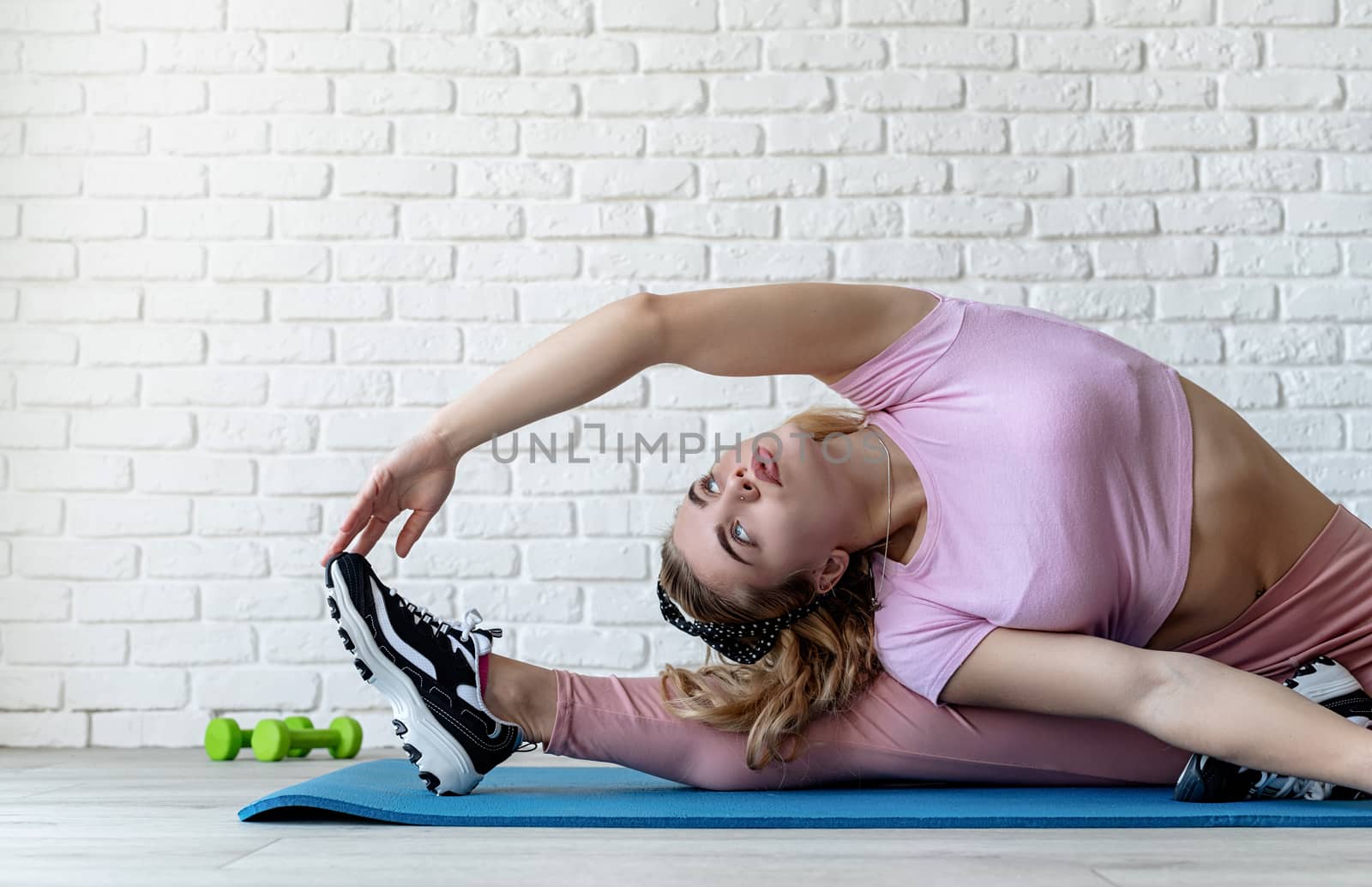 Athletic woman stretching on a fitness mat at home at white brick wall background by Desperada