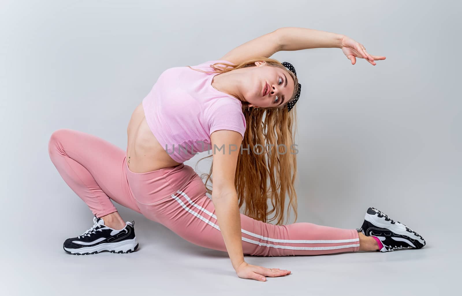 Acrobat woman wearing pink sportswear stretching in the studio isolated on gray background by Desperada