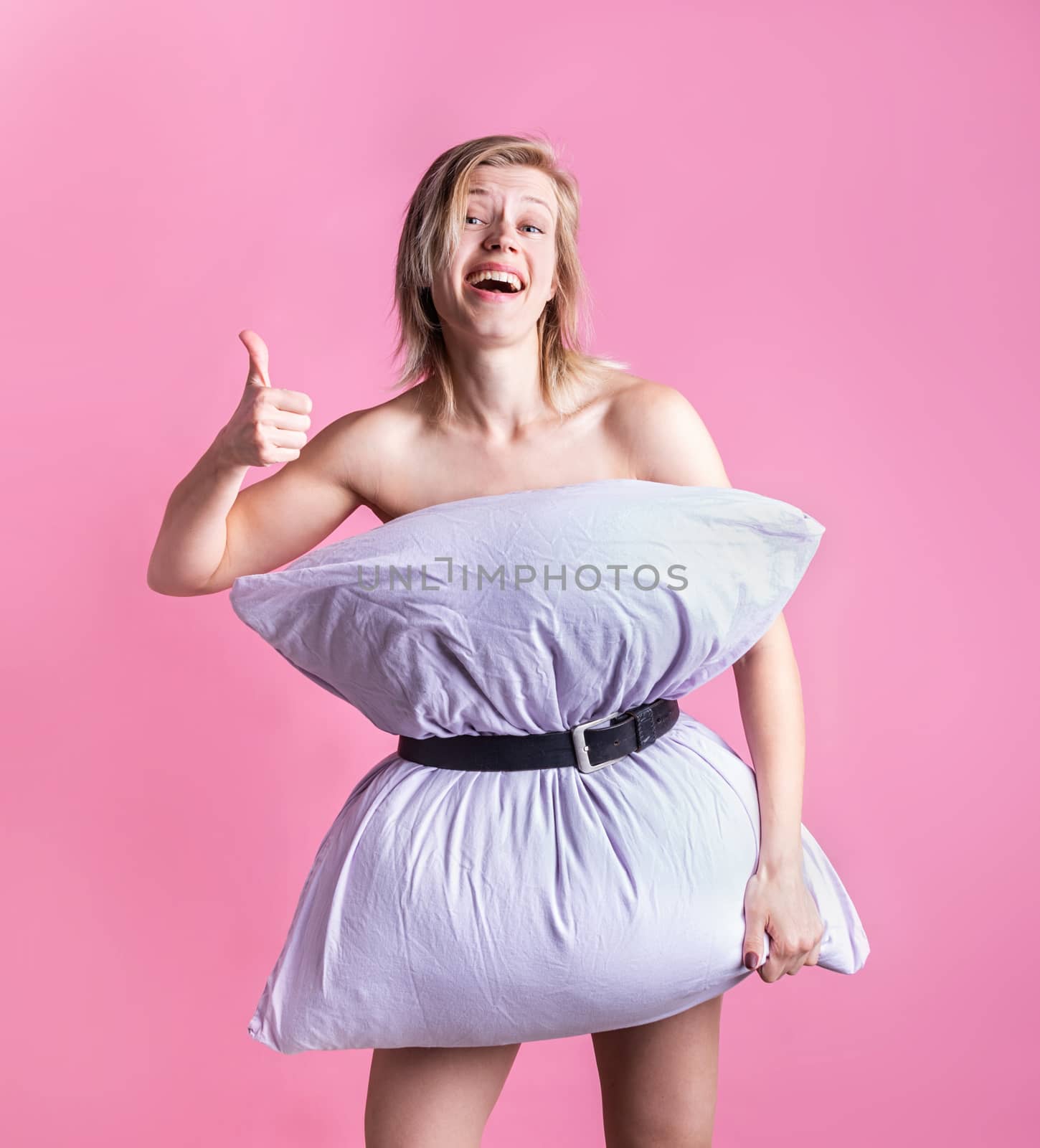 Coronavirus quarantine. Crazy quarantine. Young blond woman dressed in pillow laughing showing thumbs up on pink background. Pillow challenge outbreak