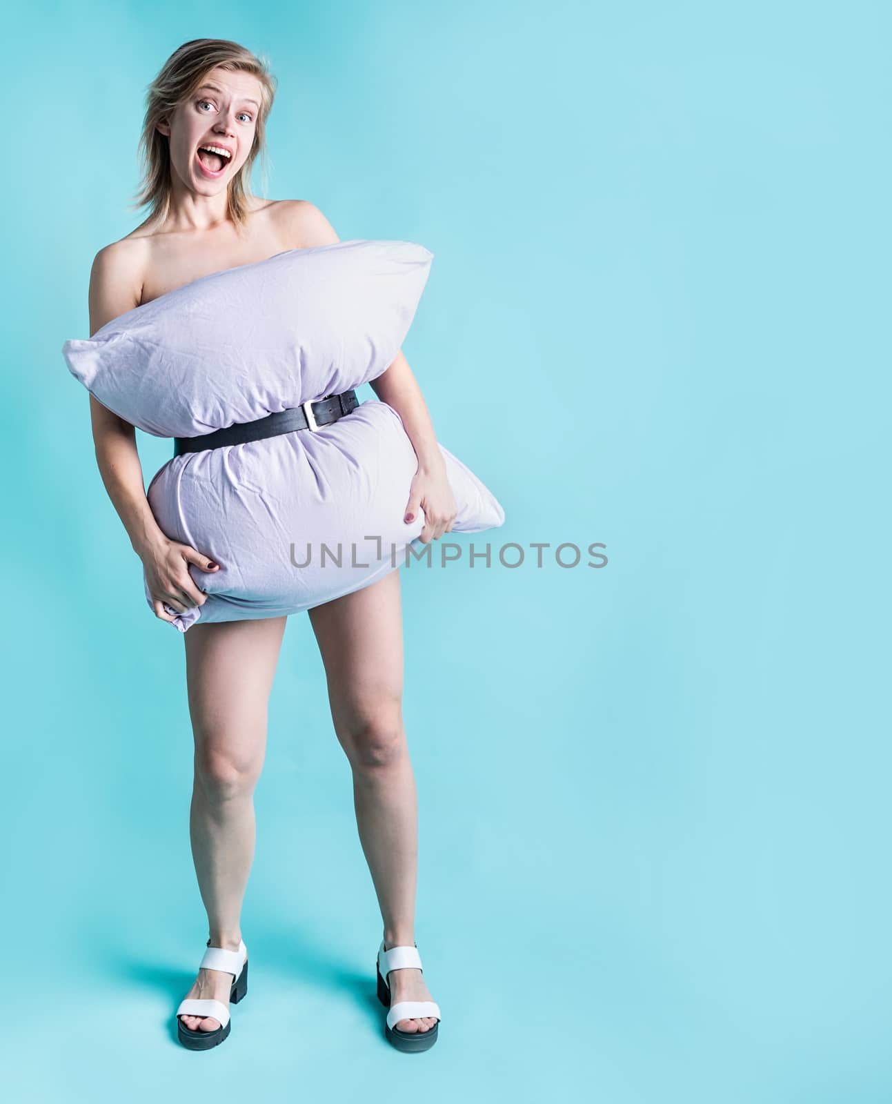 Young blond woman dressed in pillow smiling on blue background with copy space. Stay home. Crazy quarantine. Pillow challenge