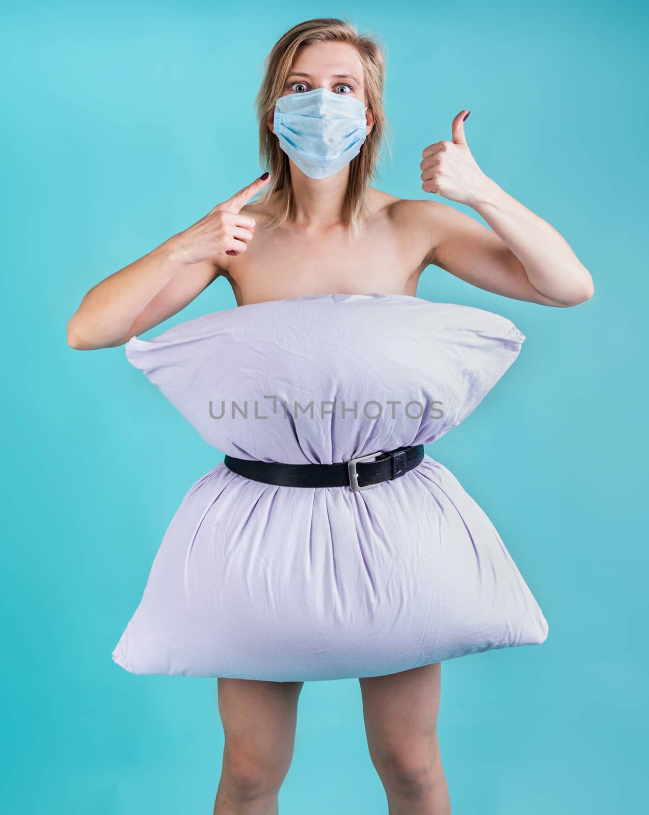 Coronavirus quarantine. Crazy quarantine. Blond woman in pillowdress wearing a mask showing thumbs up isolated on blue background. Pillow challenge outbreak