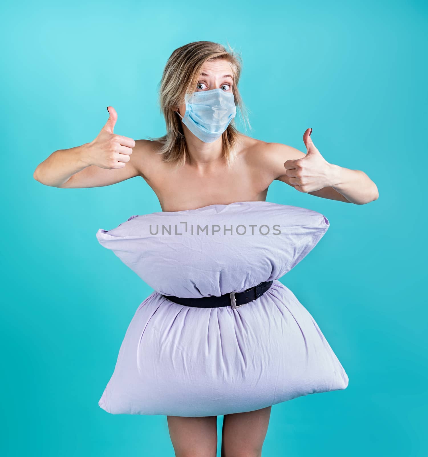 Coronavirus quarantine. Crazy quarantine. Blond woman in pillowdress wearing a mask showing thumbs up isolated on blue background. Pillow challenge outbreak