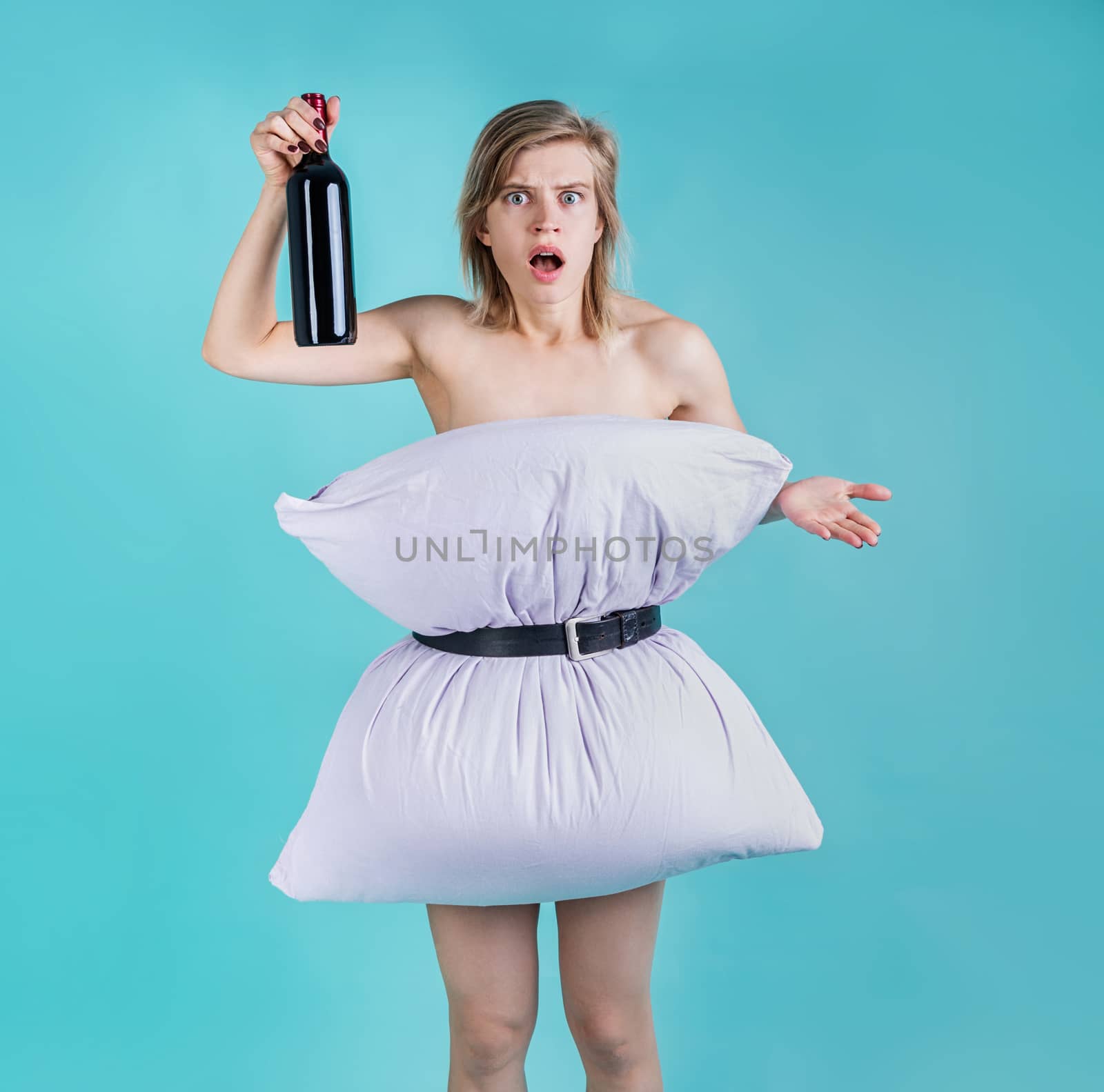Angry and surprised woman in pillowdress holding a wine bottle isolated on blue background by Desperada