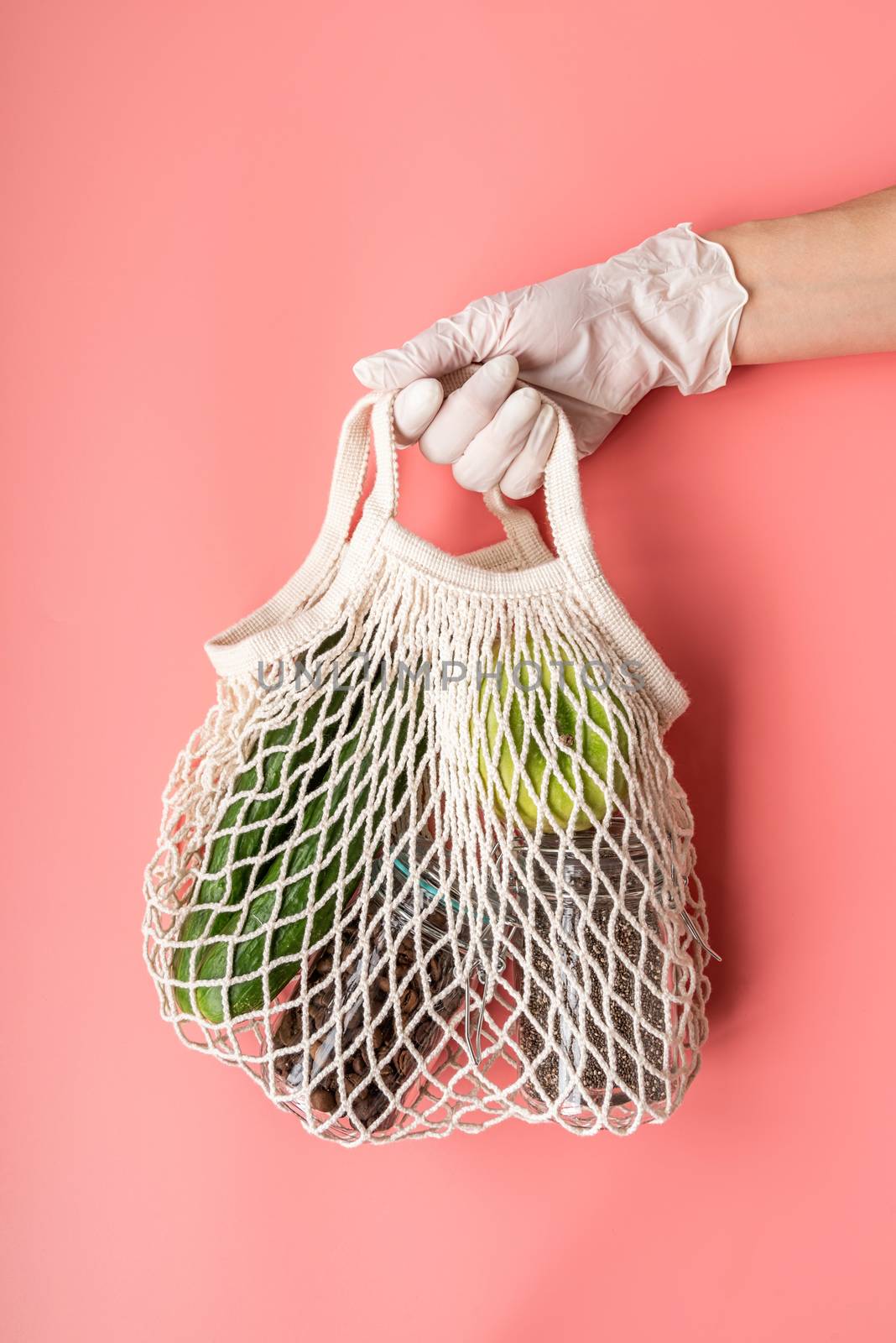 Hand in a glove holding white eco friendly mesh bag with food supplies on pink background by Desperada