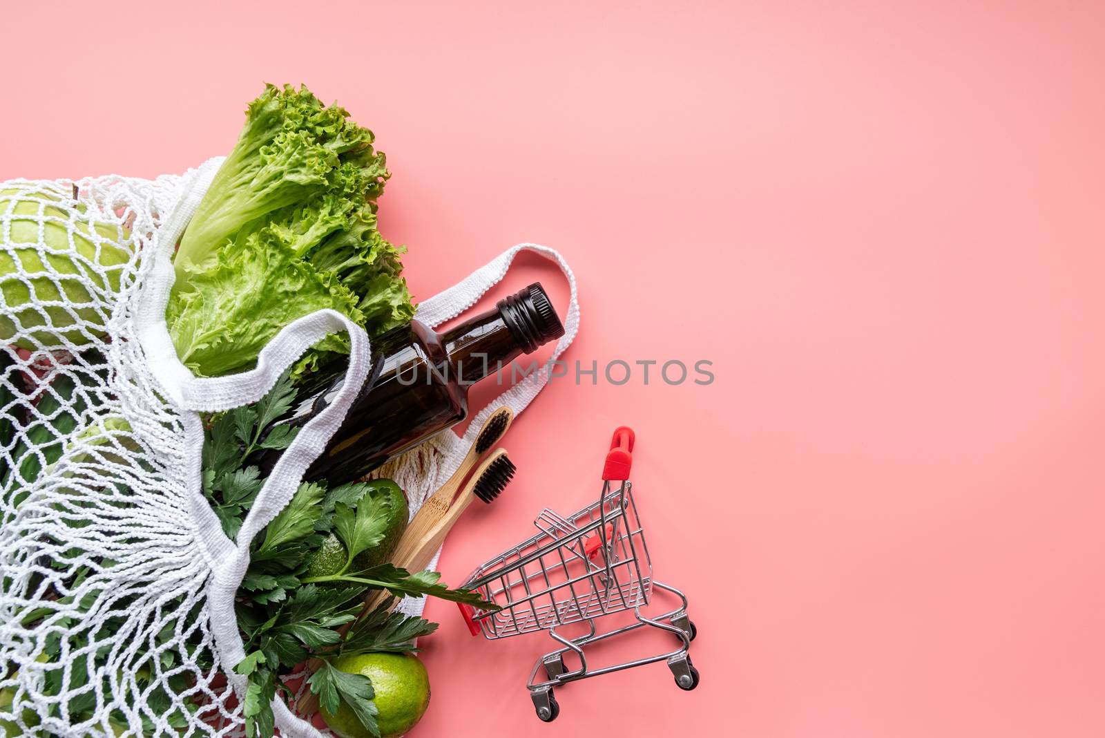Coronavirus food supplies concept. Eco friendly mesh bag with green vegetables, wooden toothbrushes, gloves and olive oil top view on pink background with copy space