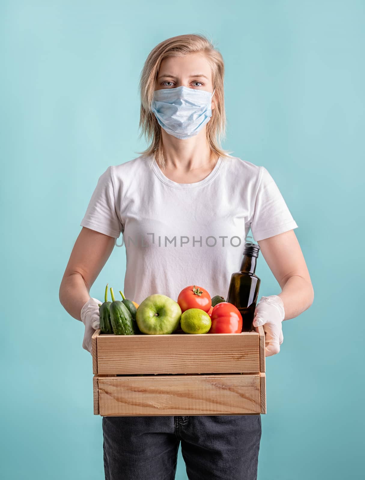 Blond woman in a mask and gloves holding a wooden box full of vegetables by Desperada