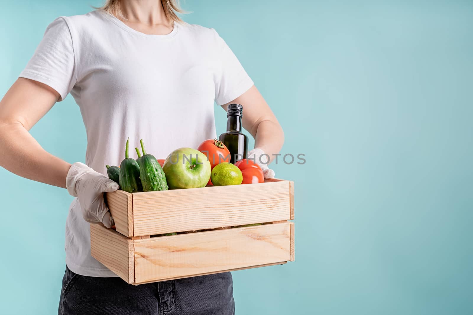 Coronavirus food supply. Woman in gloves holding a wooden box full of vegetables isolated on blue background with copy space