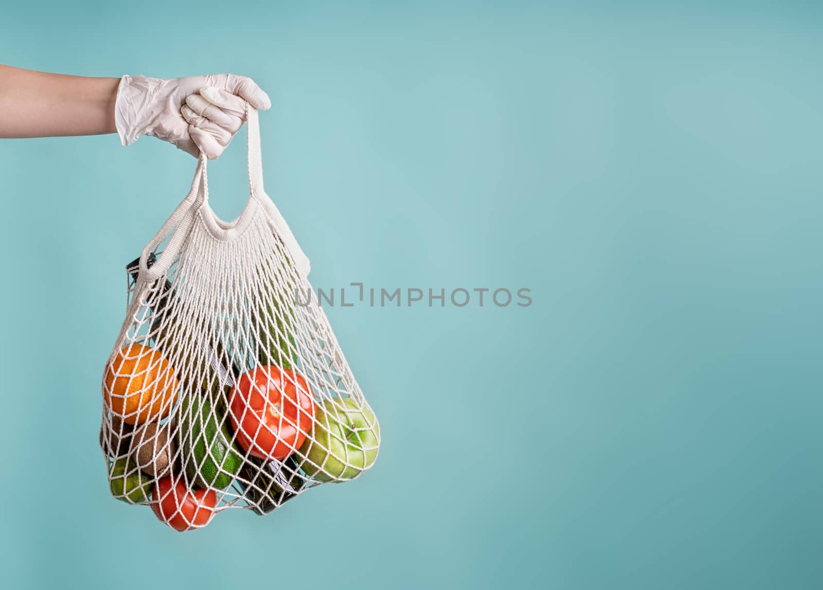 Zero waste concept. Coronavirus food delivery. Woman in glove holding a white mesh bag with vegetables isolated on blue background with copy space